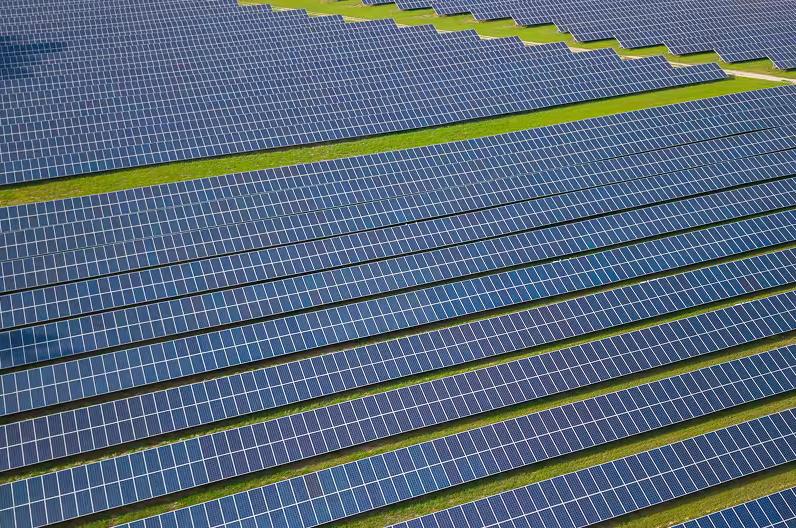 An aerial view of a large solar farm with numerous rows of blue solar panels on green grass.