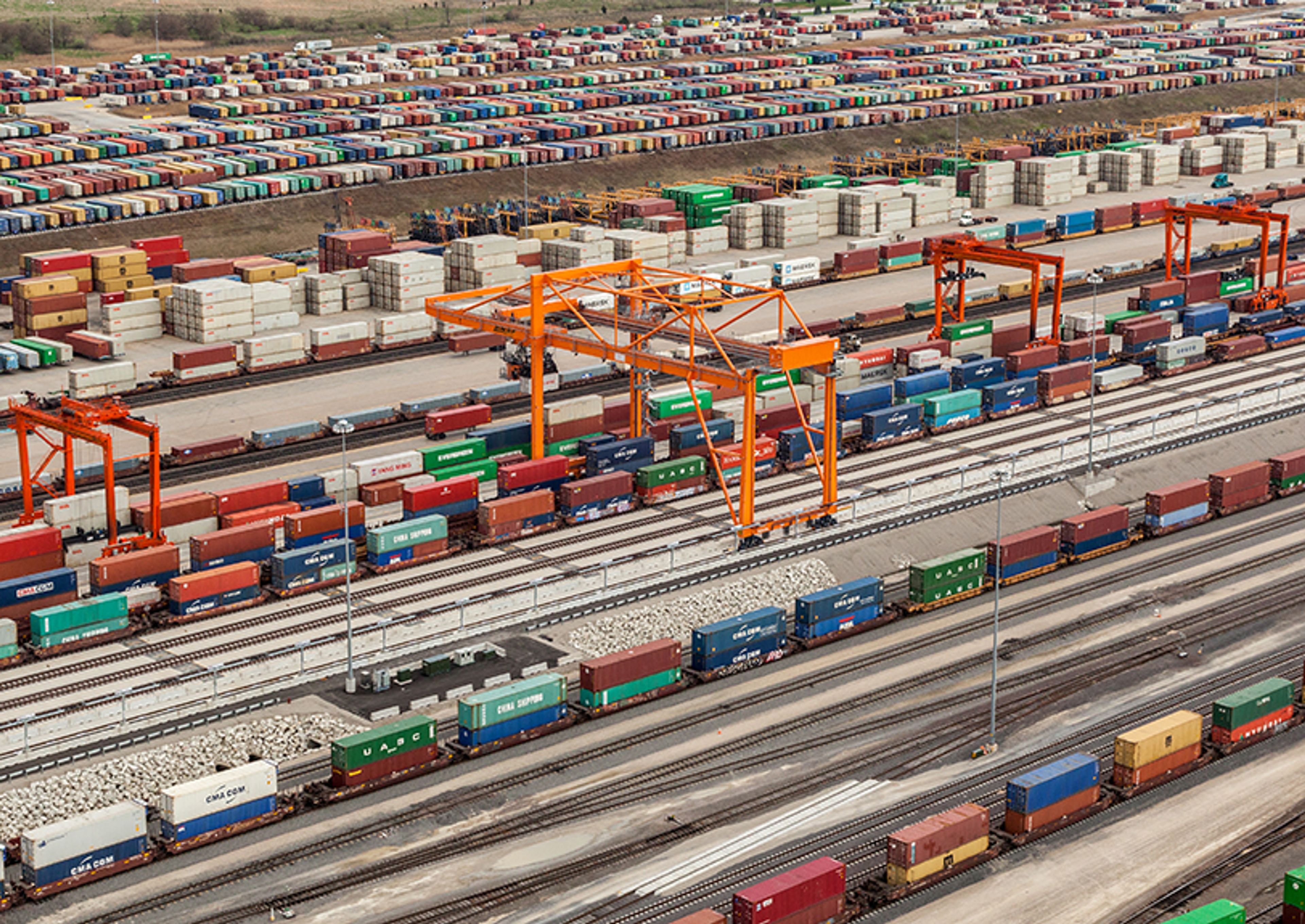 Aerial view of a BNSF intermodal yard: gantry cranes over rows of stacked shipping containers, with loaded well cars on the tracks below.