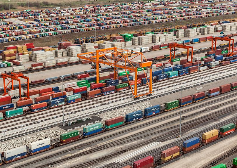 Aerial view of a BNSF intermodal yard: gantry cranes over rows of stacked shipping containers, with loaded well cars on the tracks below.
