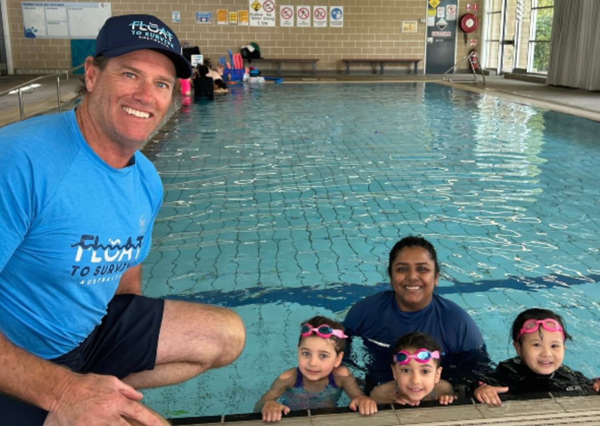 Hoppo at an indoor pool, sitting with a swim teacher with three children in the pool