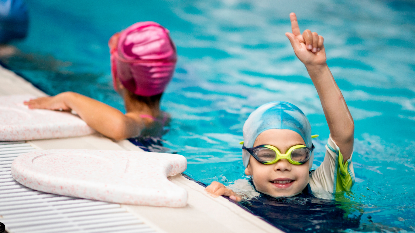 Young boy in the pool wearing goggles and a swim cap, pointing in the air and smiling at the camera