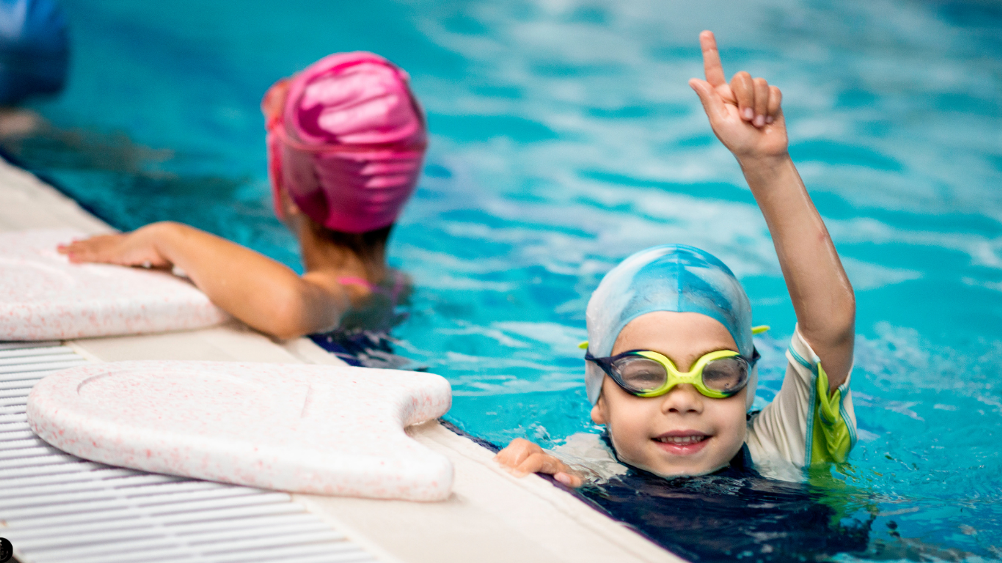 Young boy in the pool wearing goggles and a swim cap, pointing in the air and smiling at the camera 