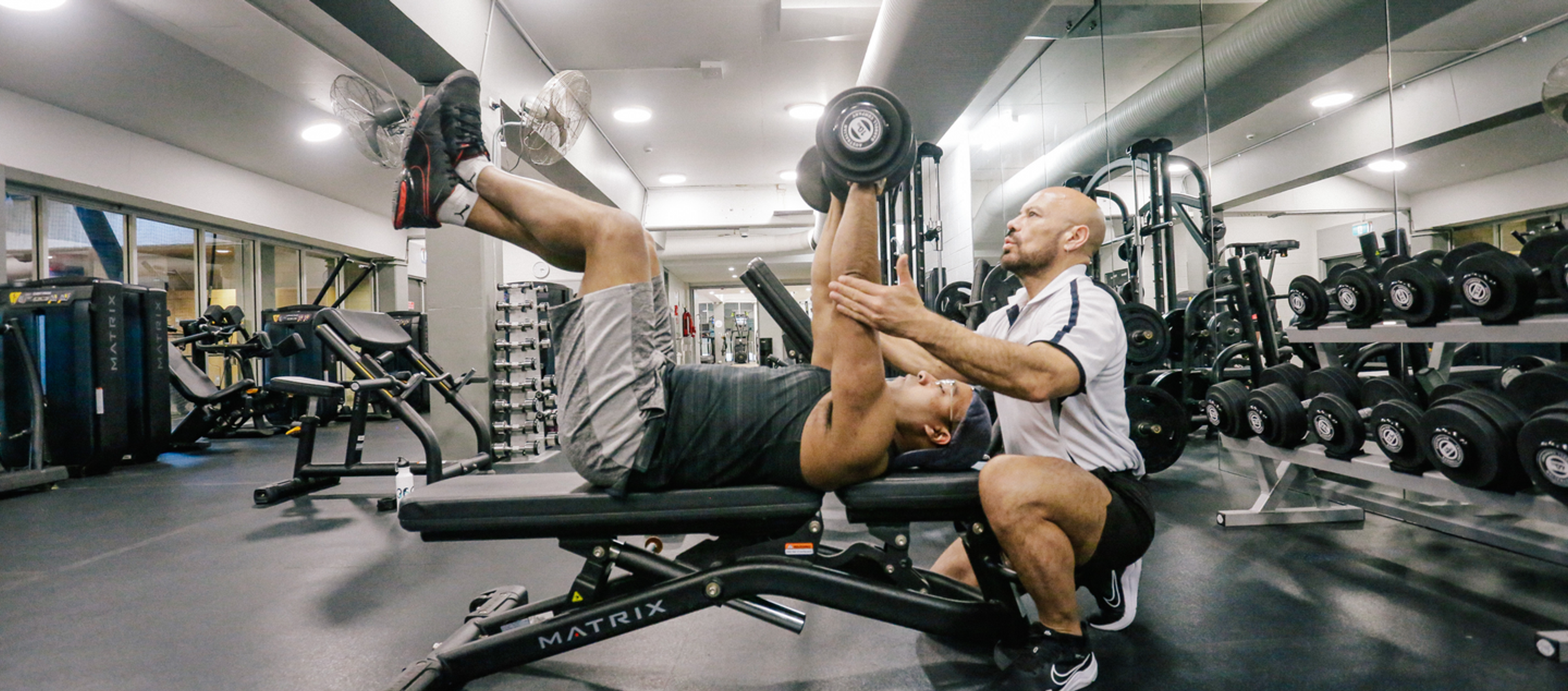 Personal trainer assists participant with dumbbell bench press in the fitness centre at Cook + Phillip Park Pool