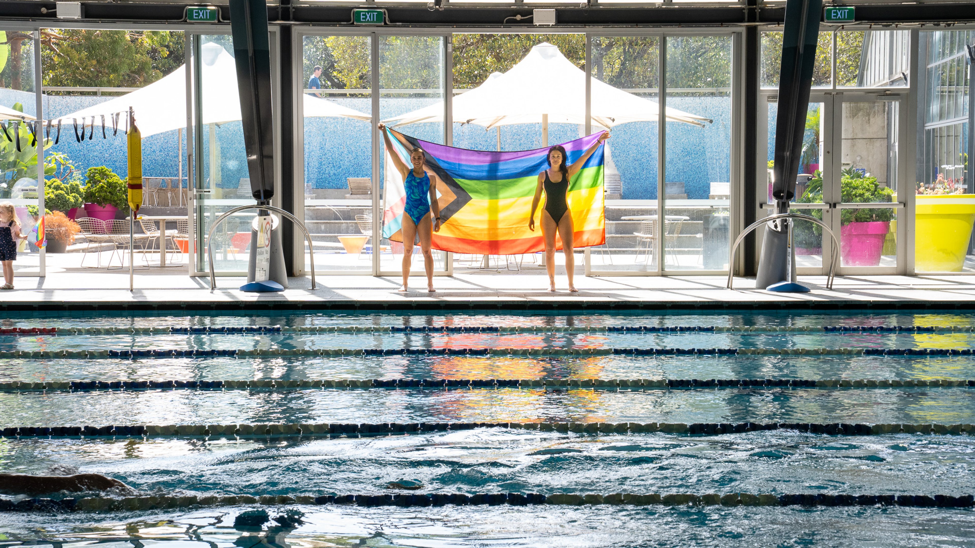 Two females holding the Pride flag next to the indoor pool at Cook + Phillip Pool