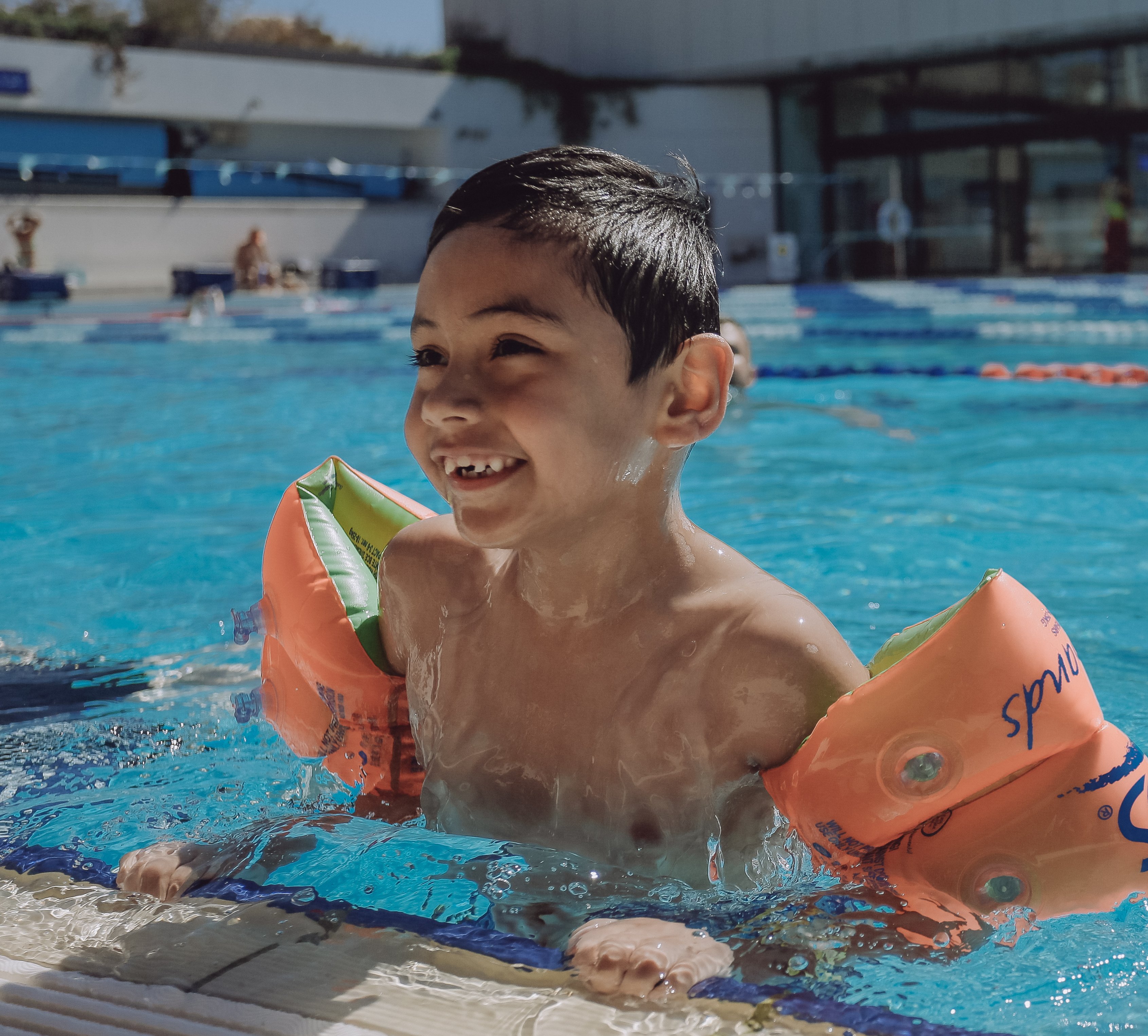Child wearing orange arm floaties smiles while holding the edge of an outdoor lap pool. Swim lanes and an aquatic centre building are visible in the background.