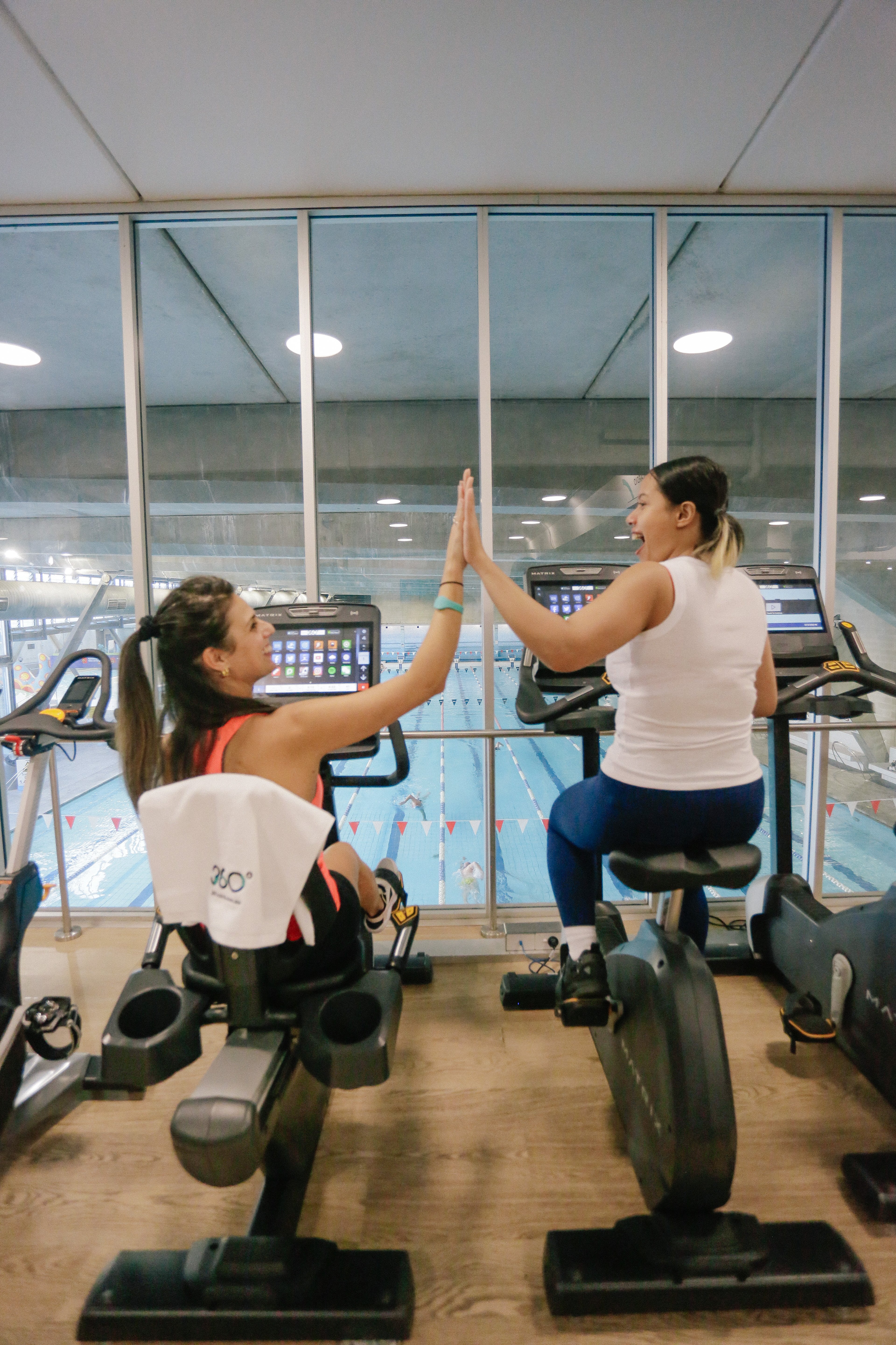 Two females giving a high five while sitting on the indoor bikes at Cook + Phillip Park Pool