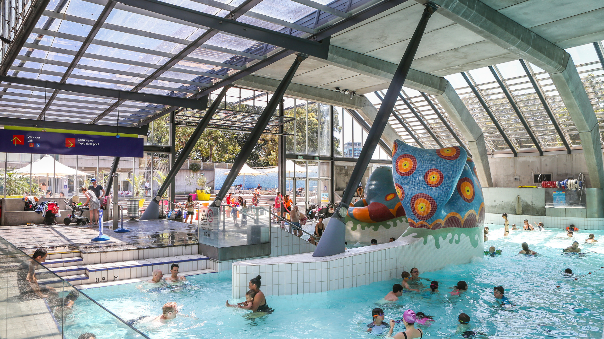 People swimming and playing in an indoor leisure pool featuring a large colourful octopus water play structure, surrounded by pool decks, seating areas and glass walls.