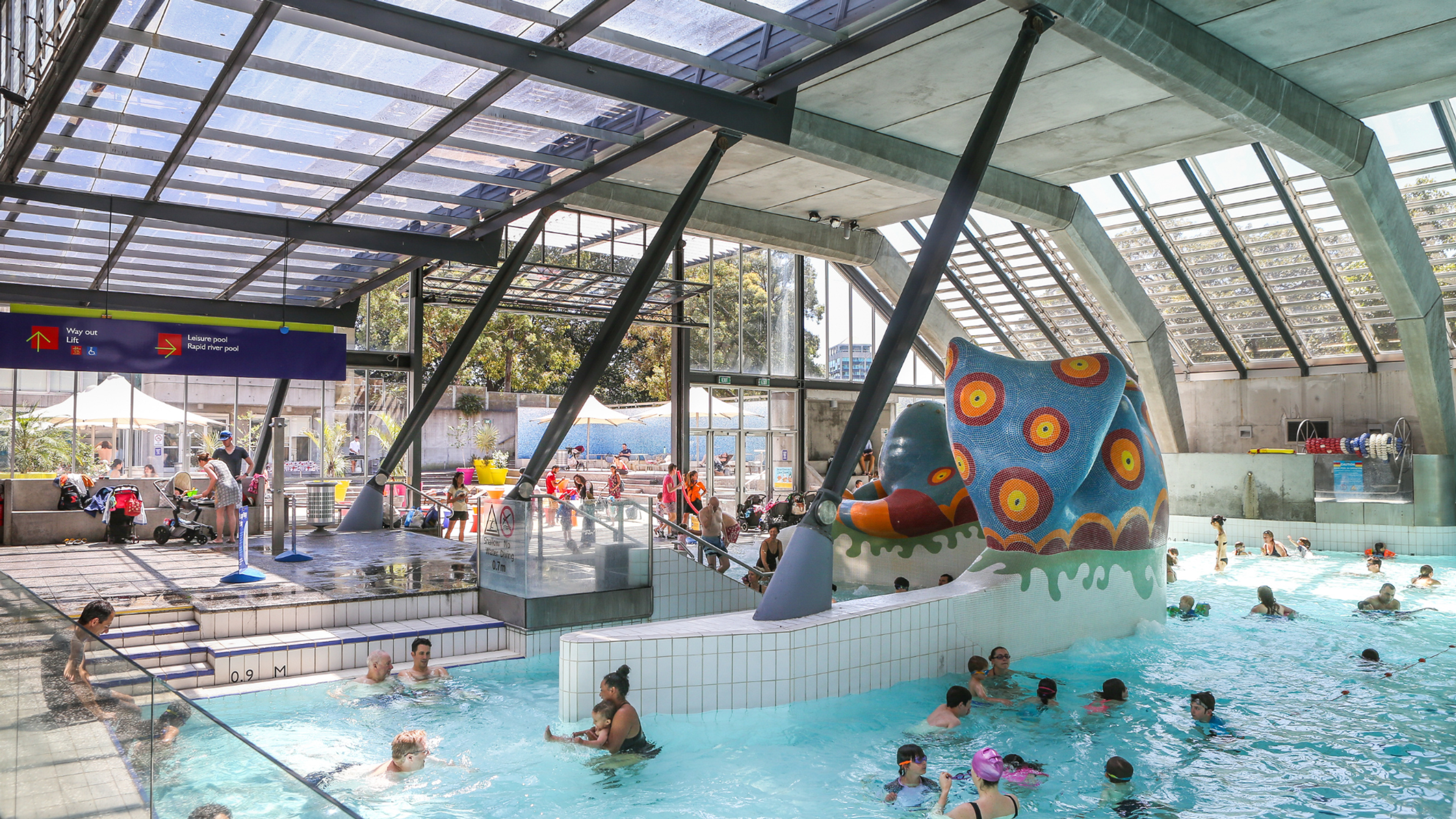 People swimming and playing in an indoor leisure pool featuring a large colourful octopus water play structure, surrounded by pool decks, seating areas and glass walls.