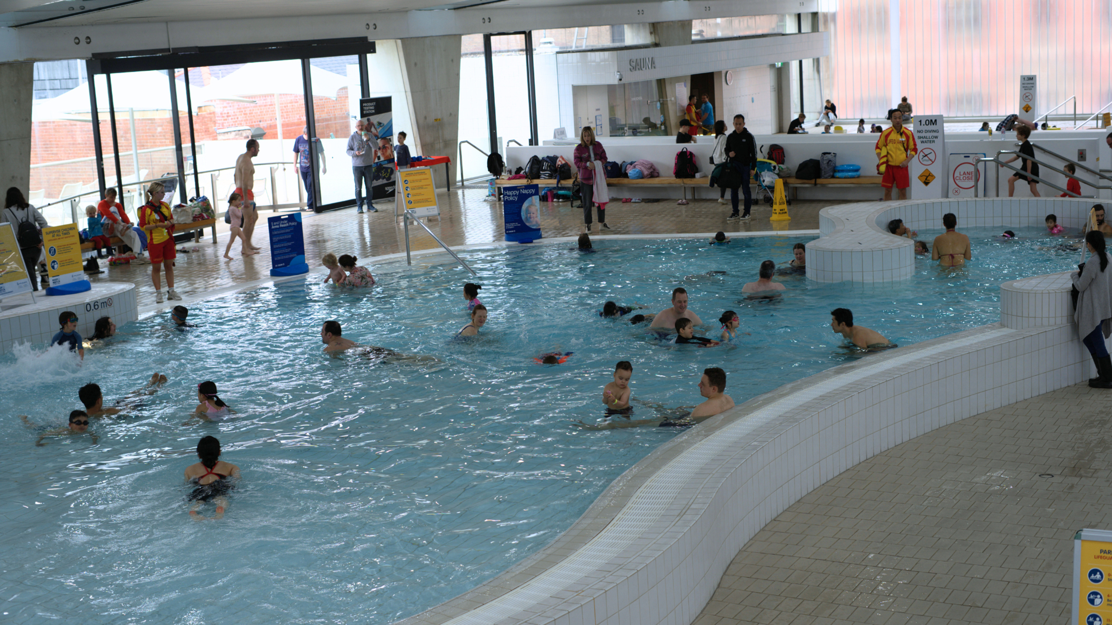 People swim and play in a large indoor leisure pool, with seating areas, poolside walkways and aquatic centre facilities visible throughout the space.