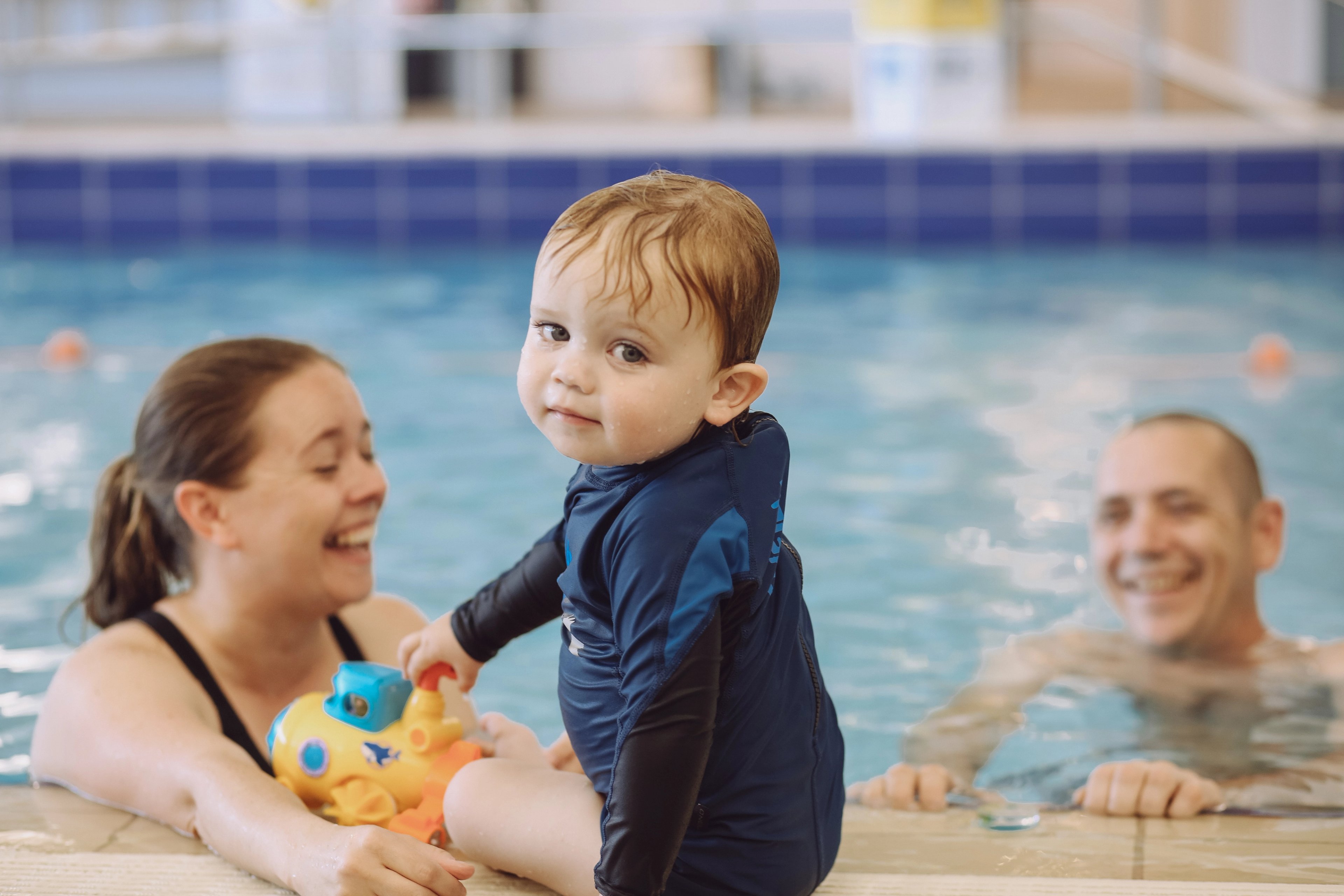 A baby sitting on the edge of the pool at Gunyama Park with his parents in the background 