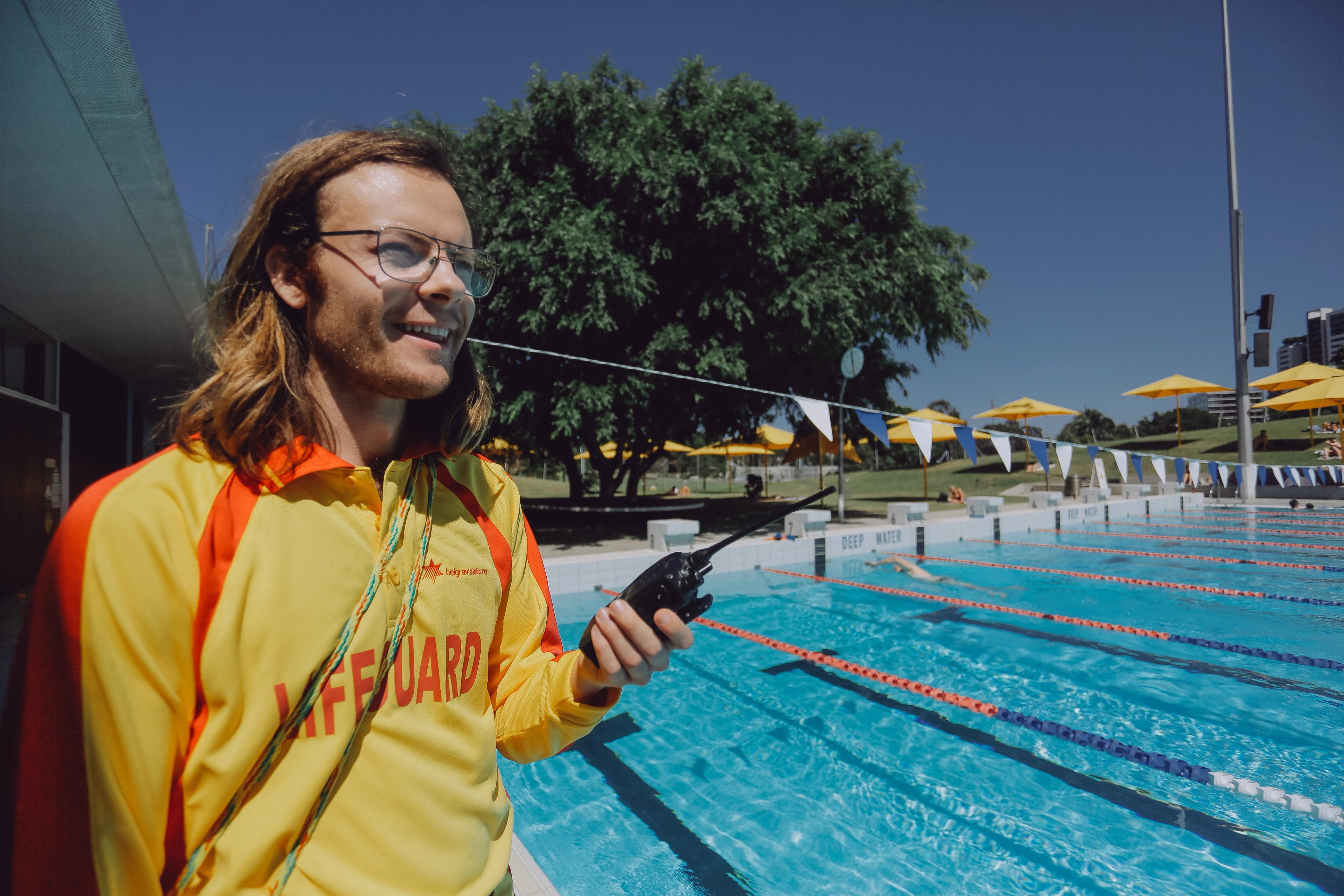Lifeguard in a yellow and red uniform stands beside an outdoor swimming pool holding a two-way radio, with marked lap lanes and shade umbrellas visible in the background.