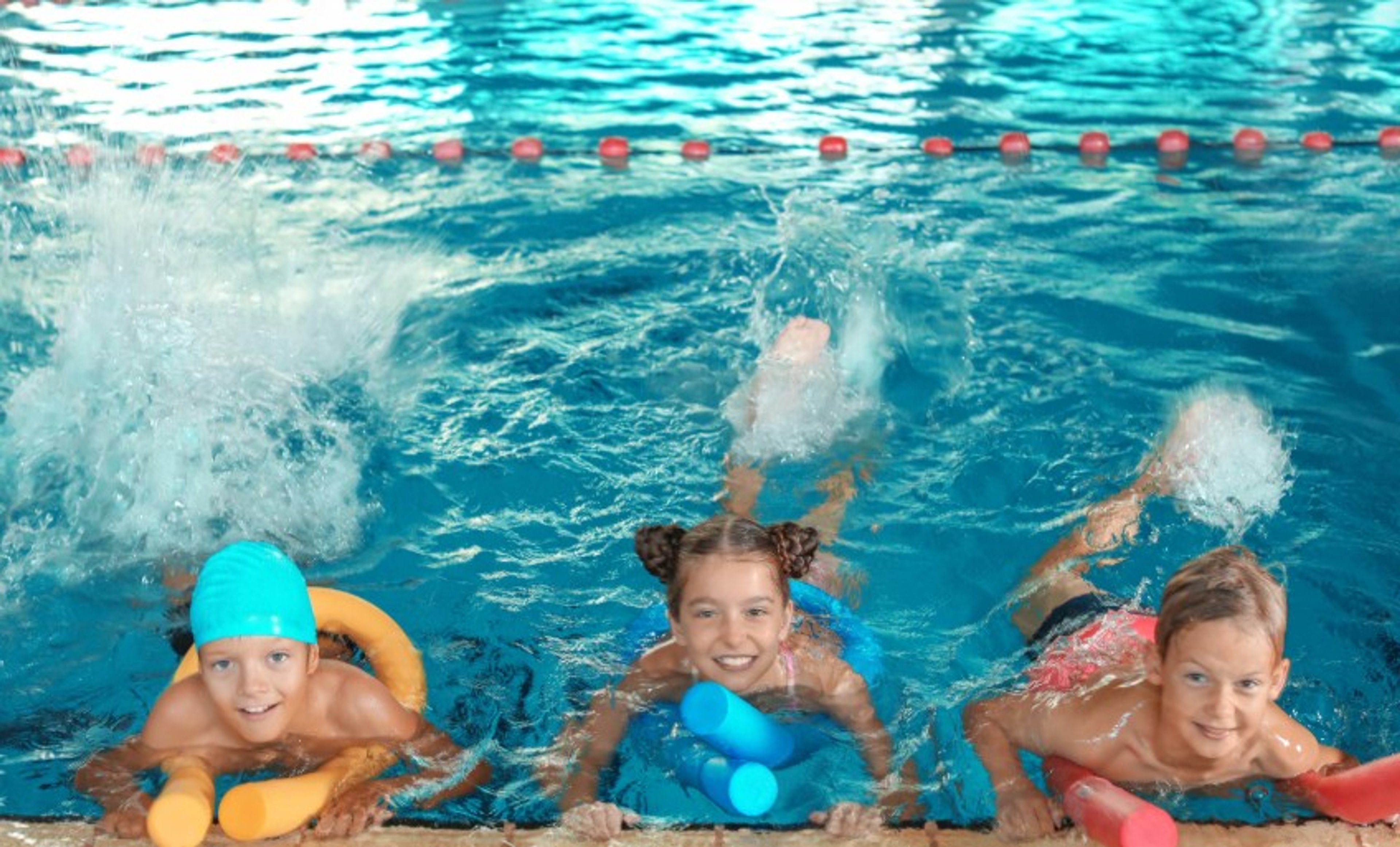 Three young kids in the pool floating on pool noodles and looking at the camera and smiling 