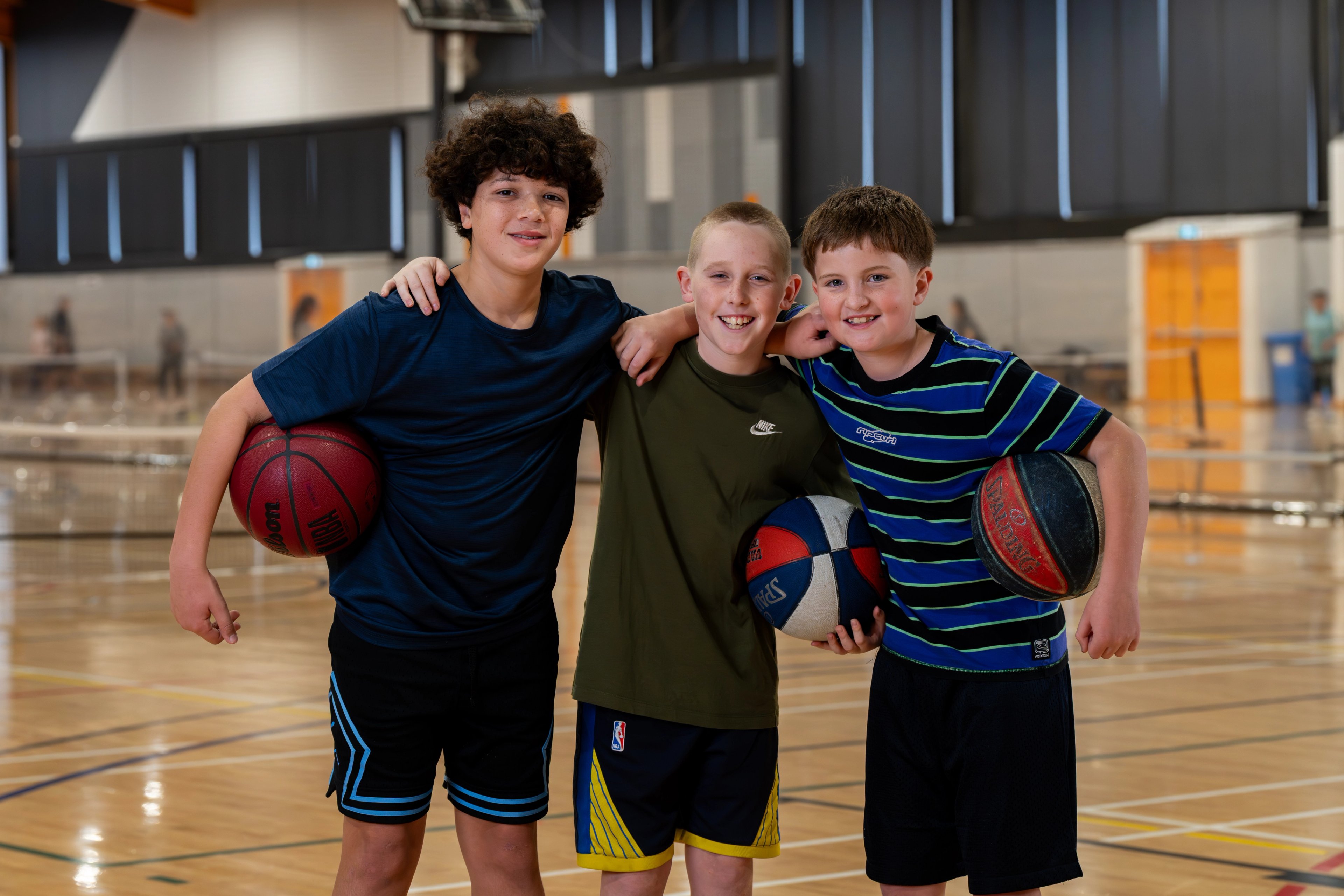 Three children standing together on an indoor basketball court, holding basketballs and wearing sports clothing. The court has a polished timber floor with marked lines, and indoor sports facilities are visible in the background.