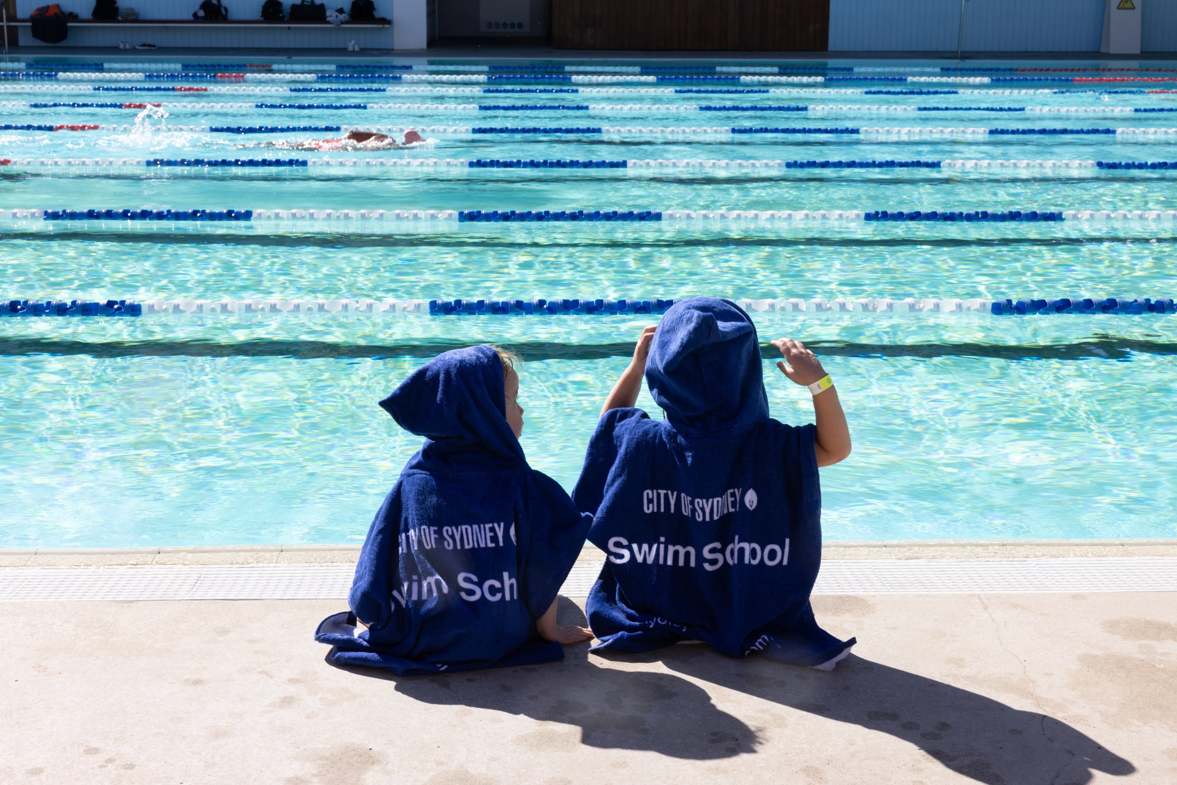 Two young children wearing swim school hooded towels and sitting by Prince Alfred Park Pool