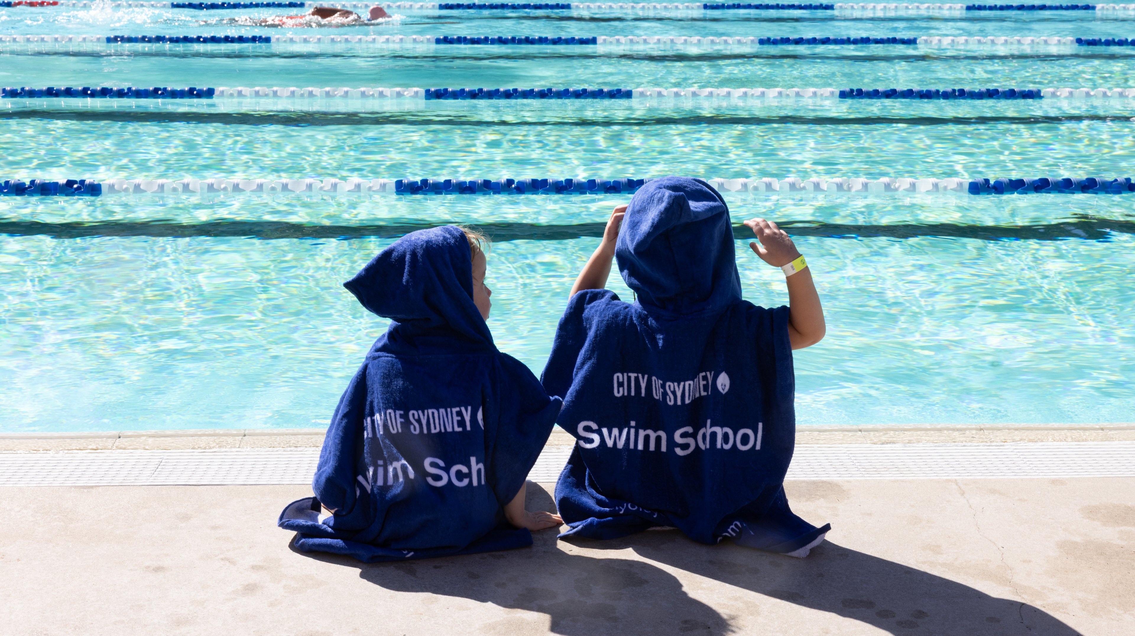 Two children sitting by the pool wearing a City of Sydney swim school hooded towel. Image: Abril Felman/City of Sydney 2025.