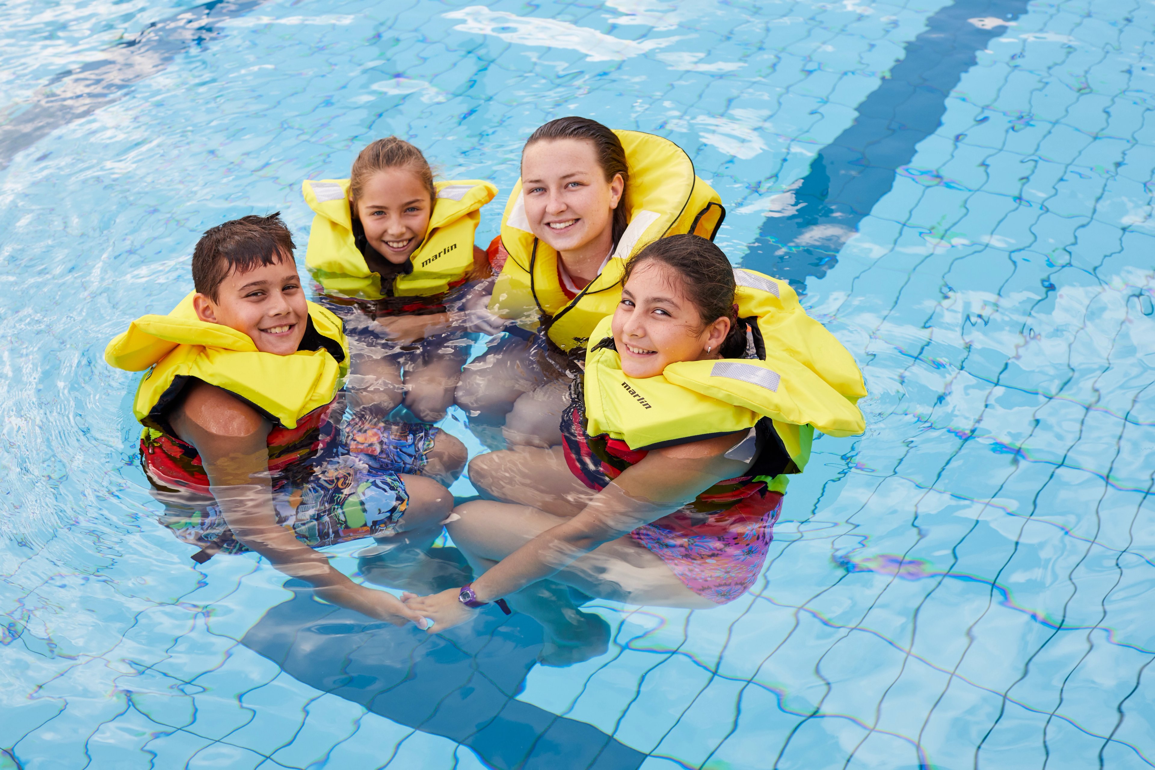 Three children and a woman floating in a pool, wearing yellow life jackets