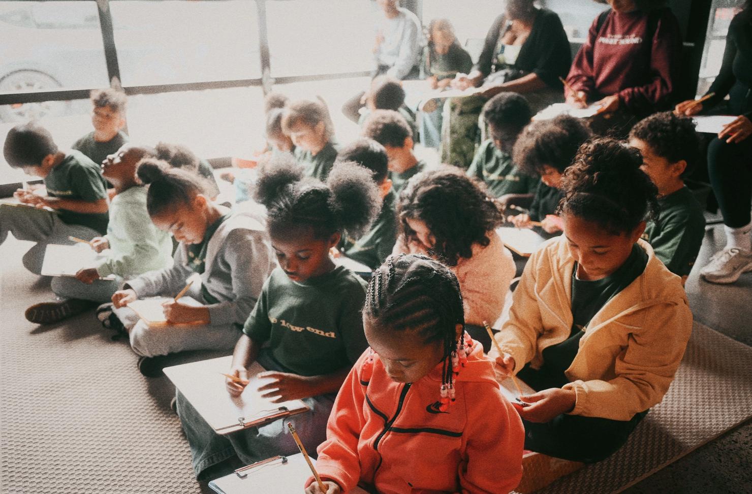 Children sitting on the floor in rows with clipboards, quietly writing.