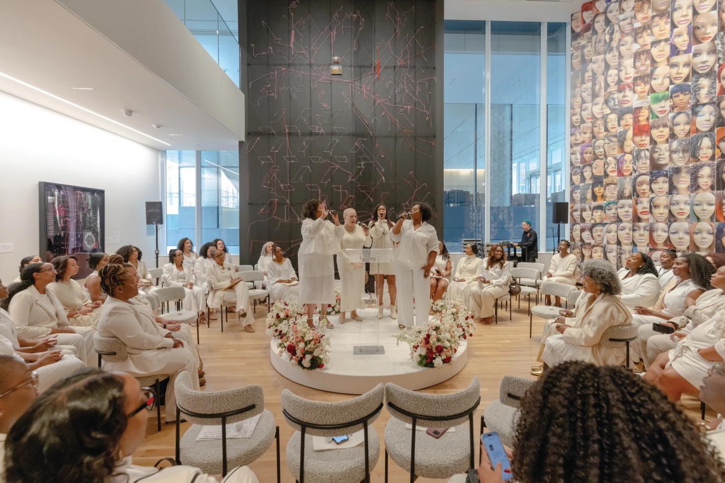 People dressed in white seated in a circle around 4 people singing on a raised platform