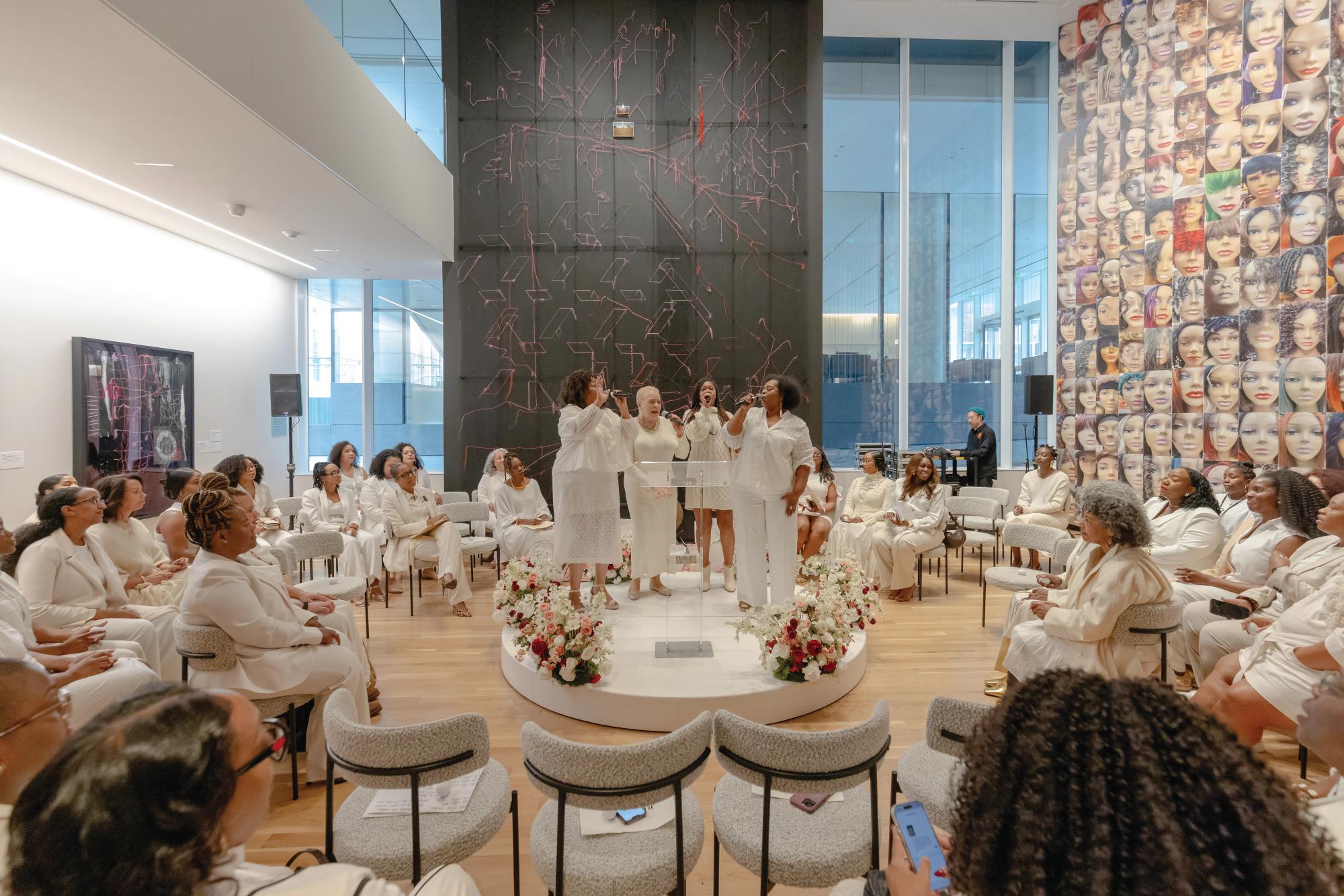 People dressed in white seated in a circle around 4 people singing on a raised platform