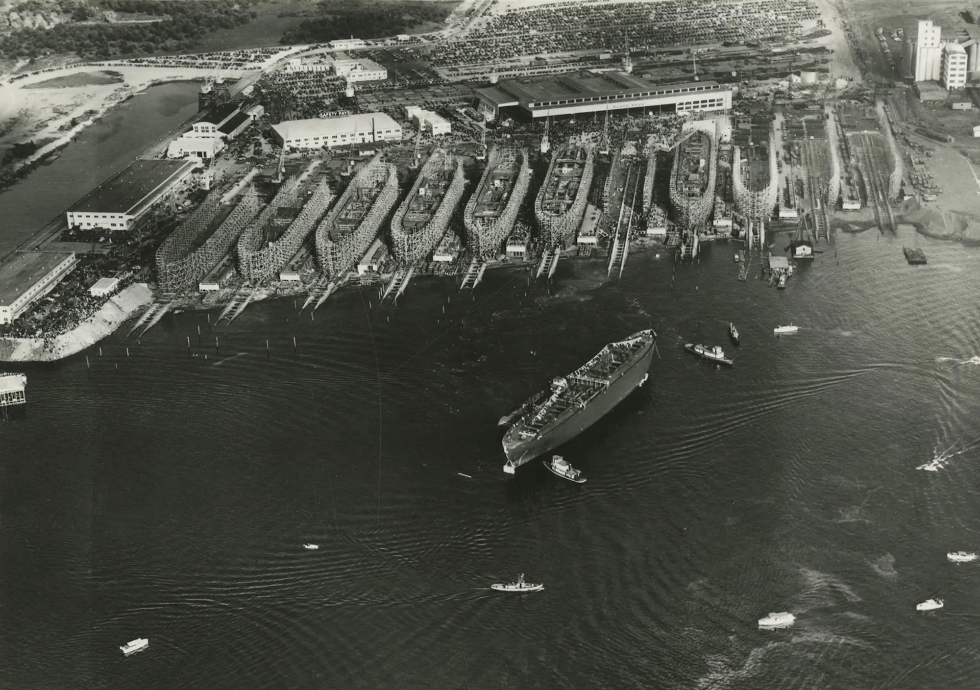 Several ships in the process of being constructed on the shore, one ship floating in the river.