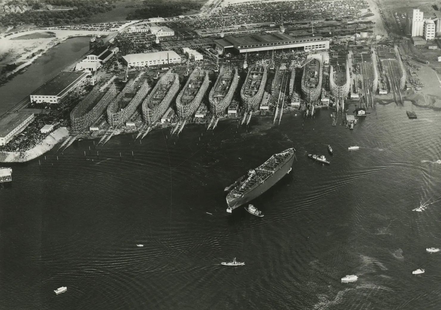 Several ships in the process of being constructed on the shore, one ship floating in the river.