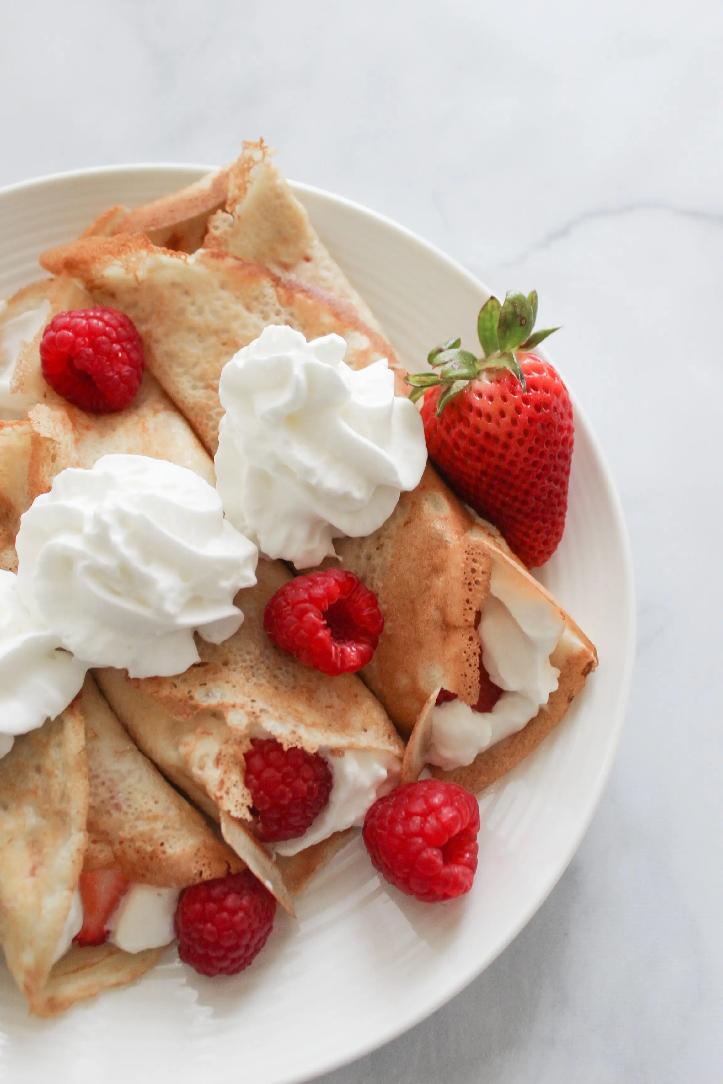 three rolled crepes with fruit, butter and whipped cream in the centres. The tops have 1 dollops of whipped cream each and a large strawberry sits on the side of the plate.