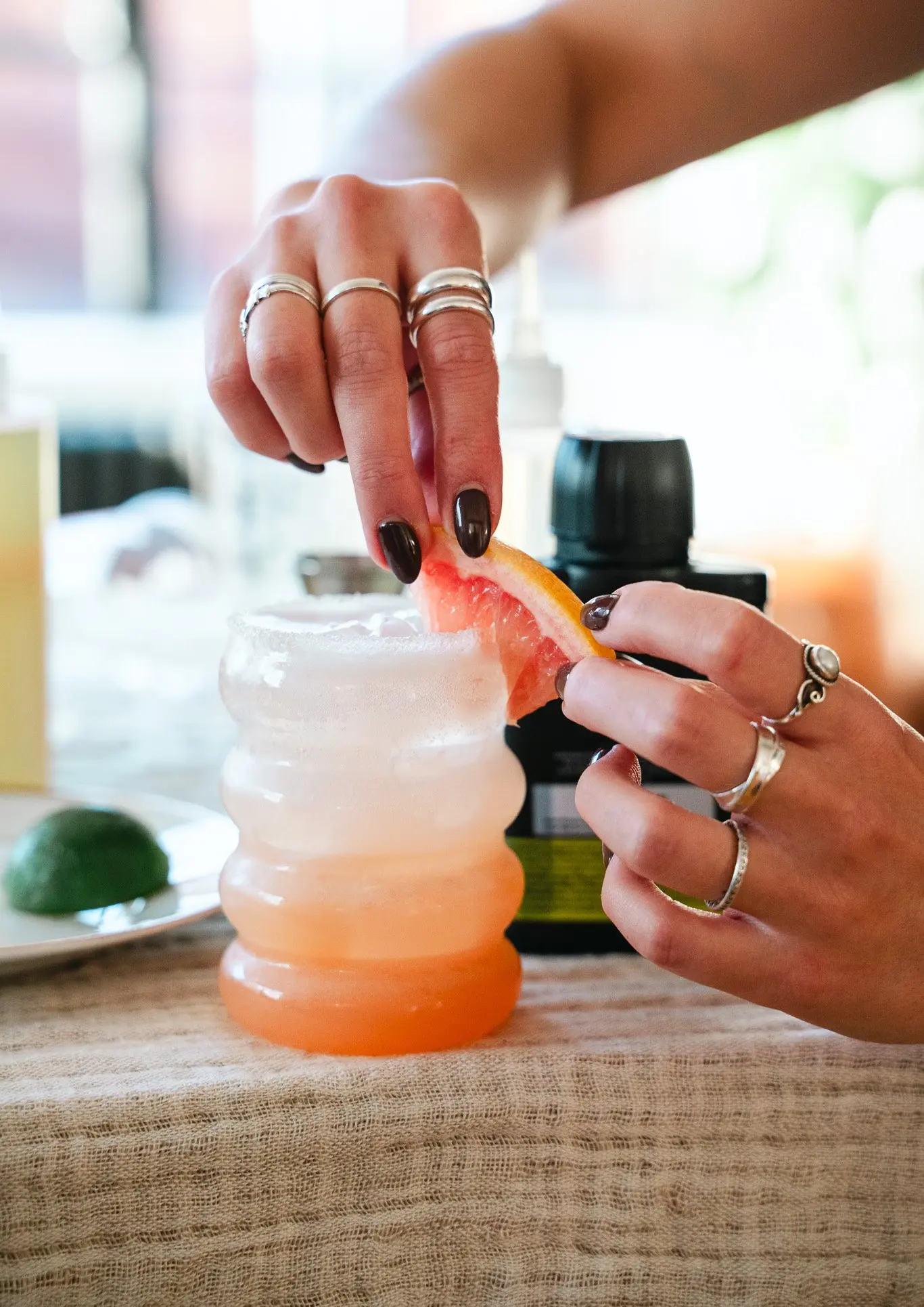 A bubble glass full of a pink and white layered Paloma mocktail, with a hand adding a slice of grapefruit to the side of the glass.