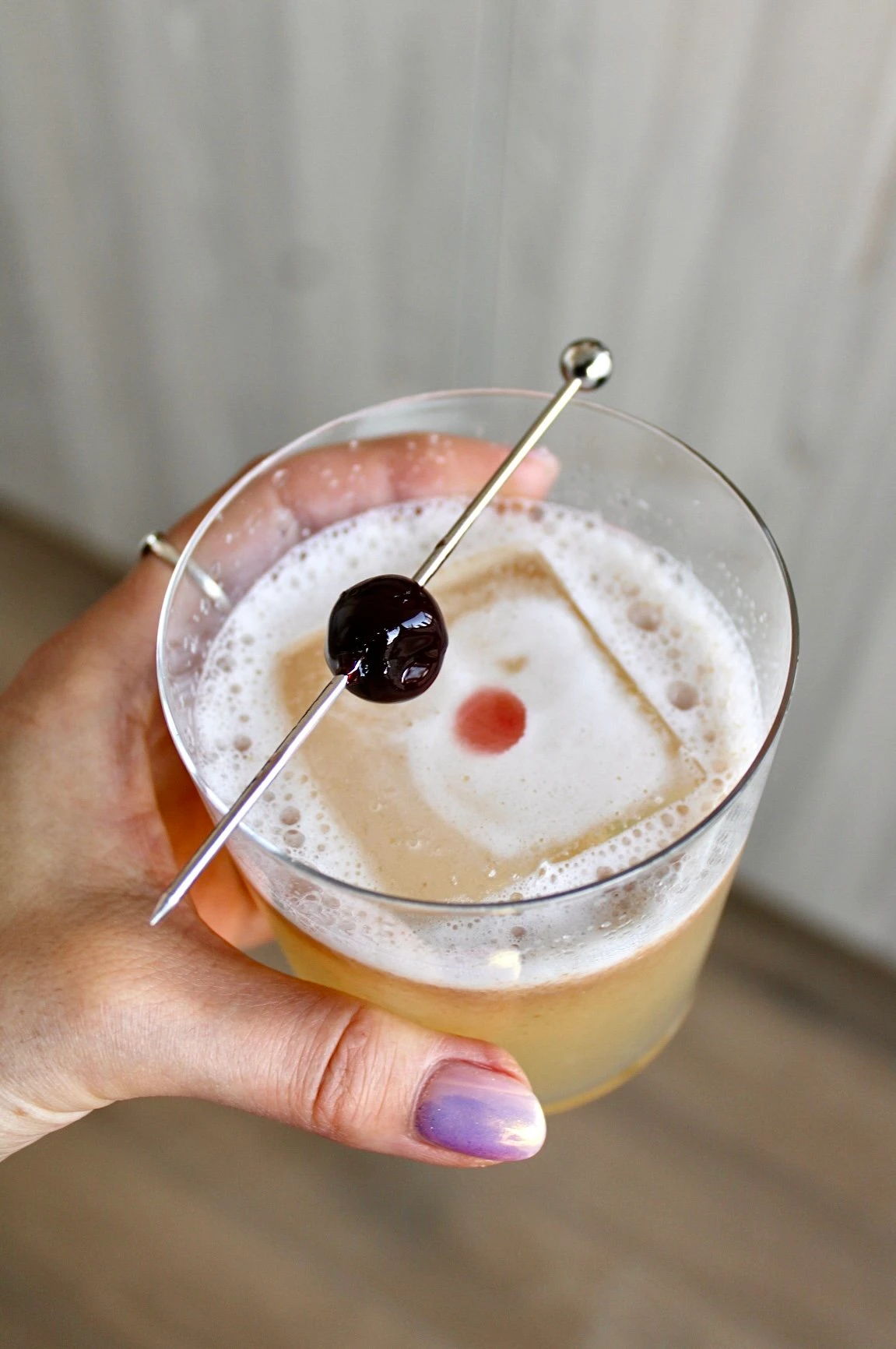 A rocks glass being held by a hand with purple nail polish, inside of which is an infused old fashioned mocktail with a singular ice cube.