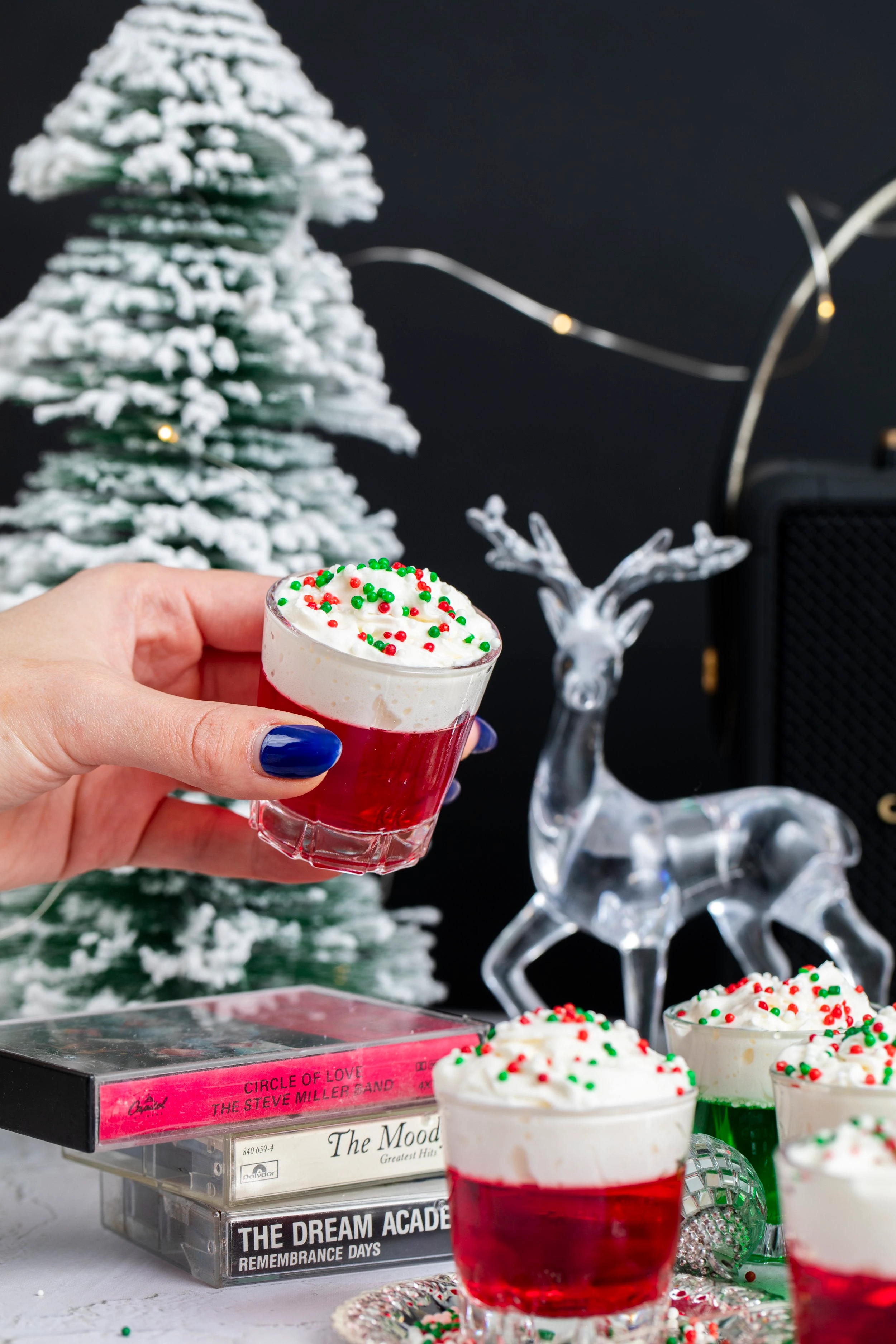 A hand picking up a red jello shot topped with whipped cream as three other jello shots rest next to it on the table. The shots are red and green and the decor surrounding them are Christmas themed.