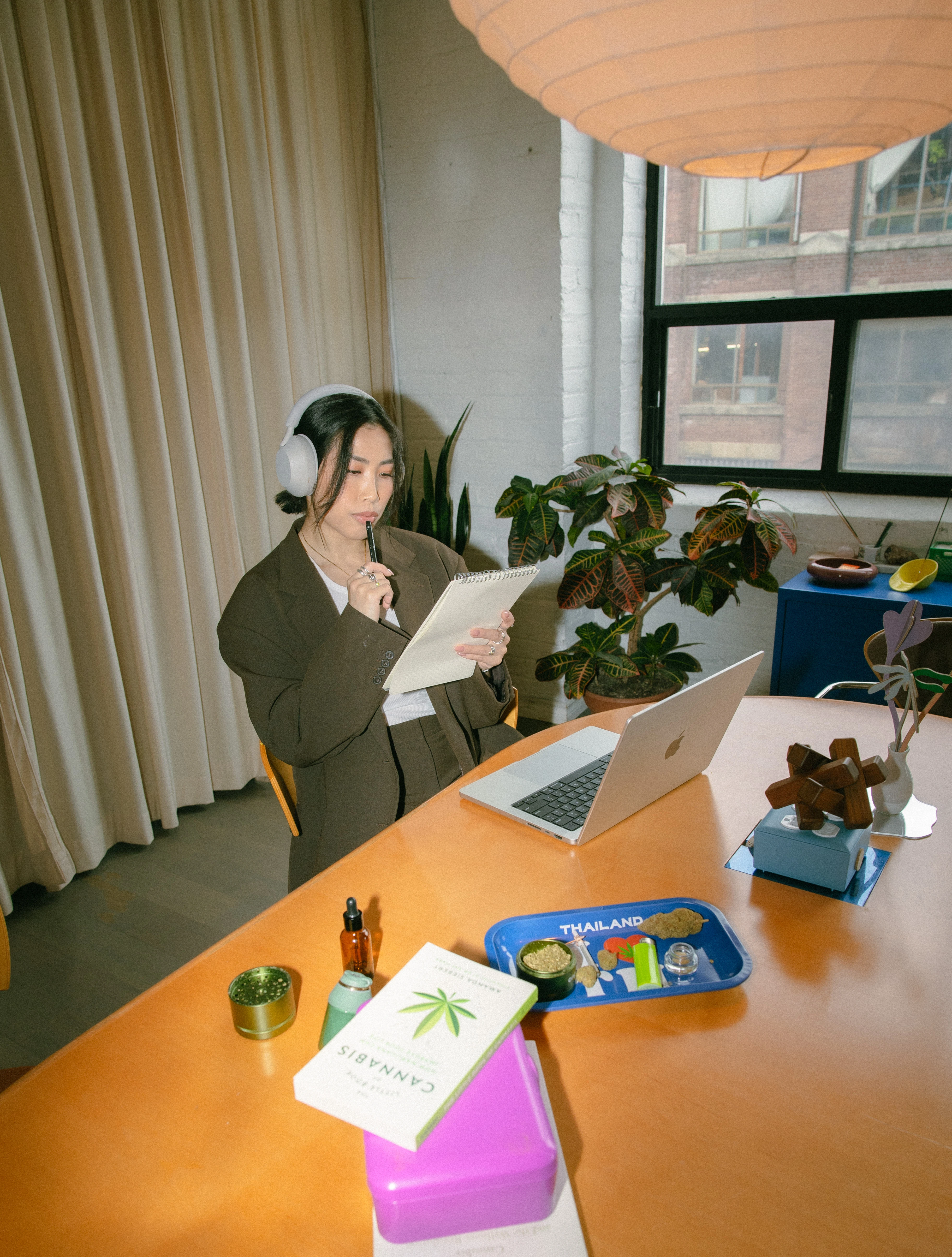 Anna The Cannabinista looking at a notepad she's holding, with her computer on the brown table below her. She wears over the ear headphones on her head.