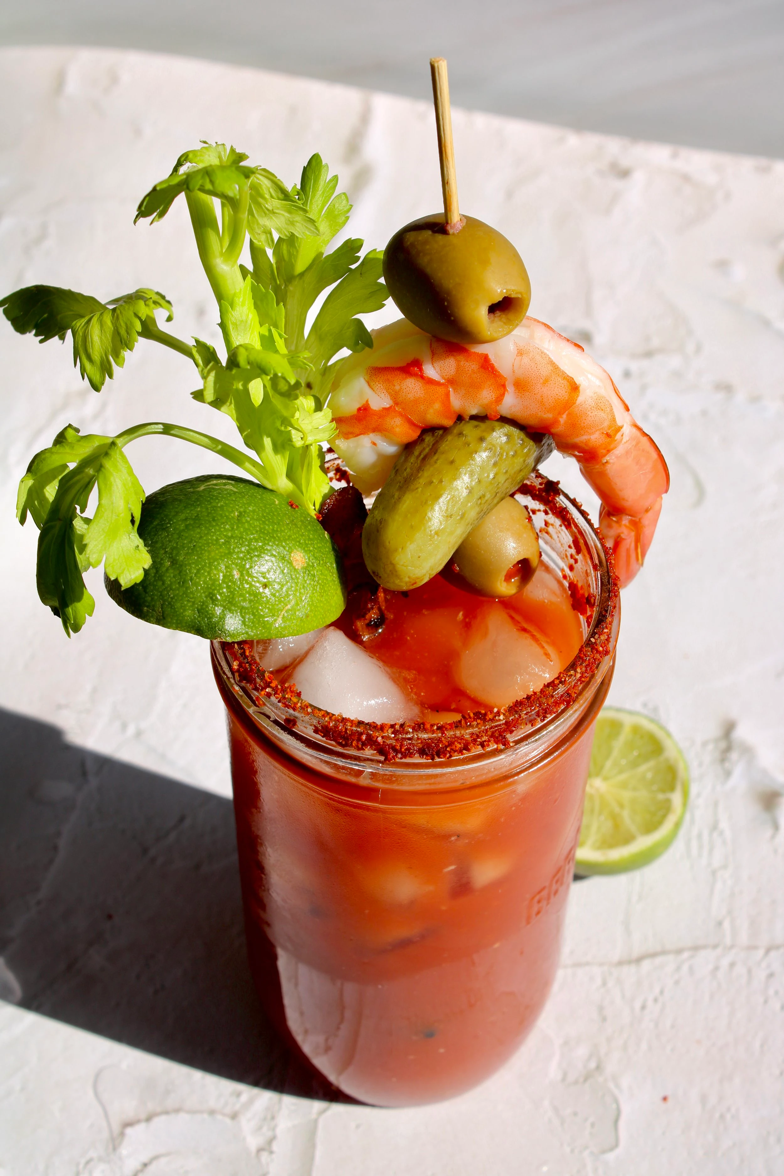 A jar full of an infused Caesar mocktail garnished with a variety of pickled snacks as well as a shrimp, stick of celery and lime.