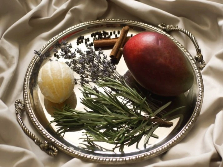 A silver tray resting on a satin sheet which holds a few sprigs of rosemary, a peeled clementine orange, a mango, some dried lavender and two cinnamon sticks. 