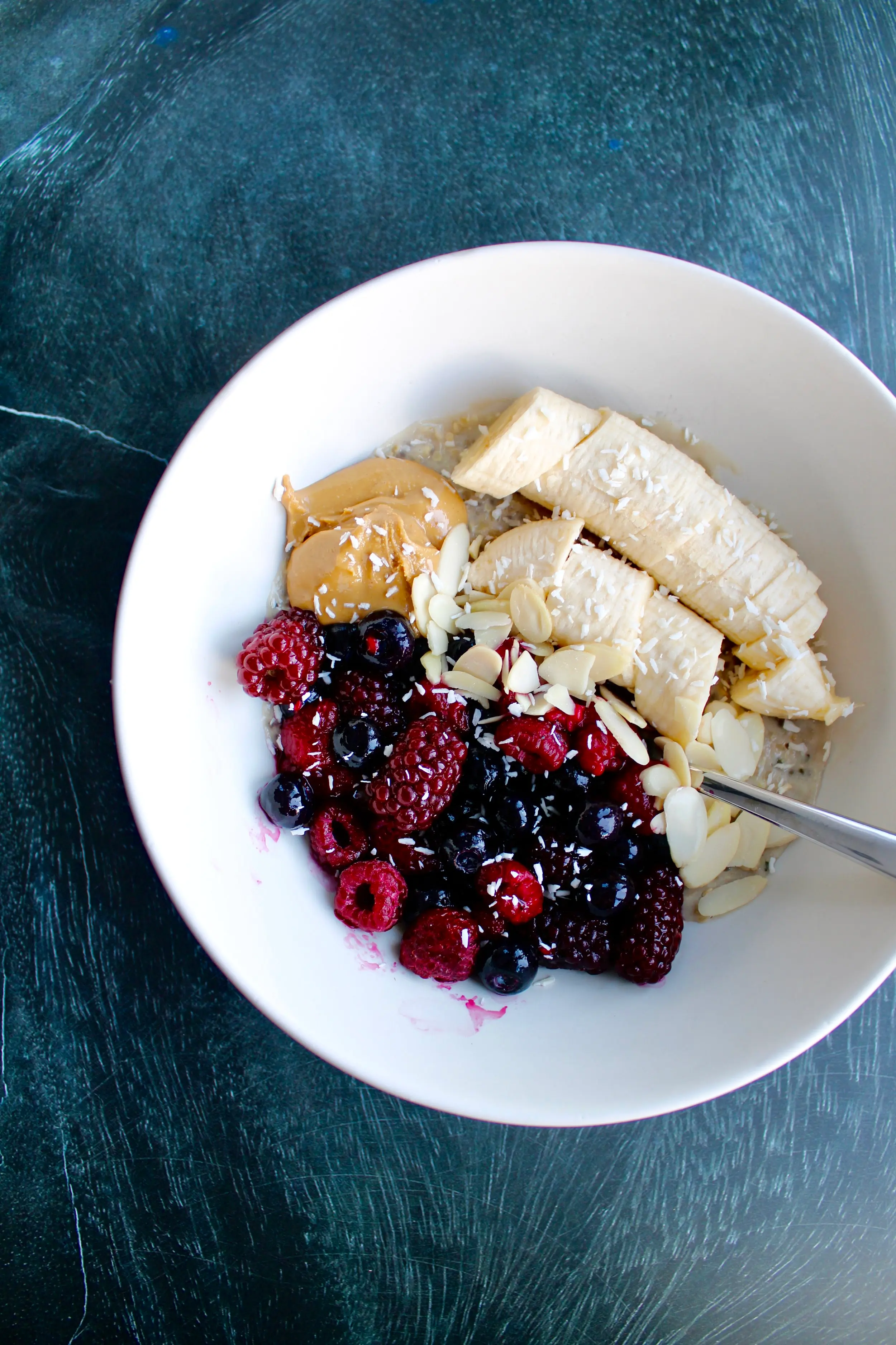 a white bowl of cooked, rolled oats with berries on the left side as well as a chopped up banana on the top right. There is also a dollop of peanut butter on the top left side of the bowl.