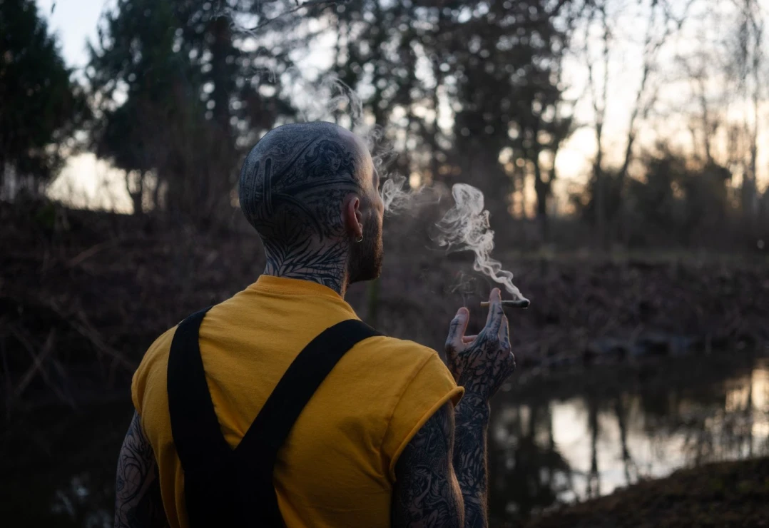 tattooed bald man in a yellow t-shirt smoking a joint in a forest at sunset