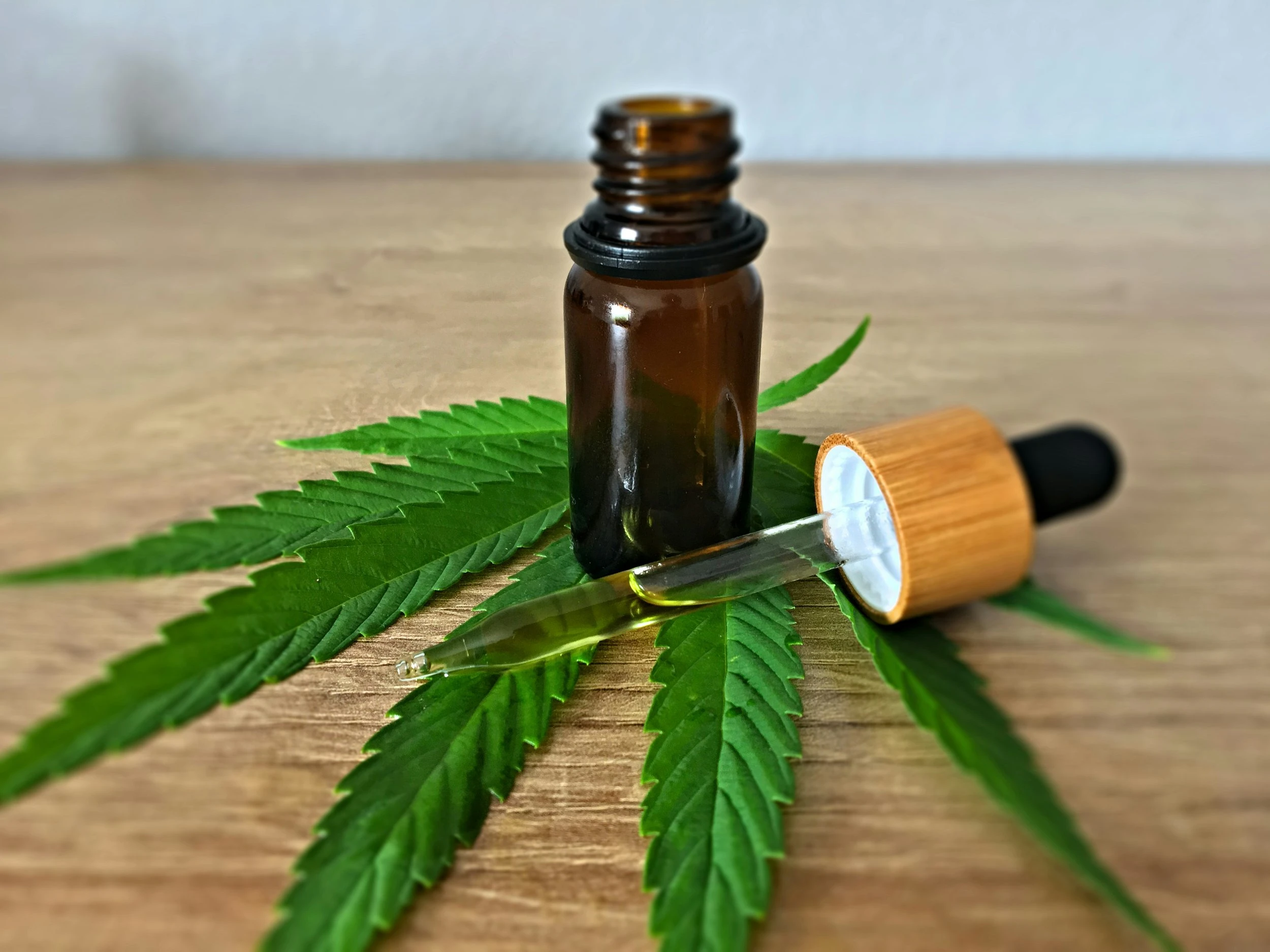 A green cannabis leaf resting on a wooden table with a bottle and its corresponding dropper resting on top of the leaf.