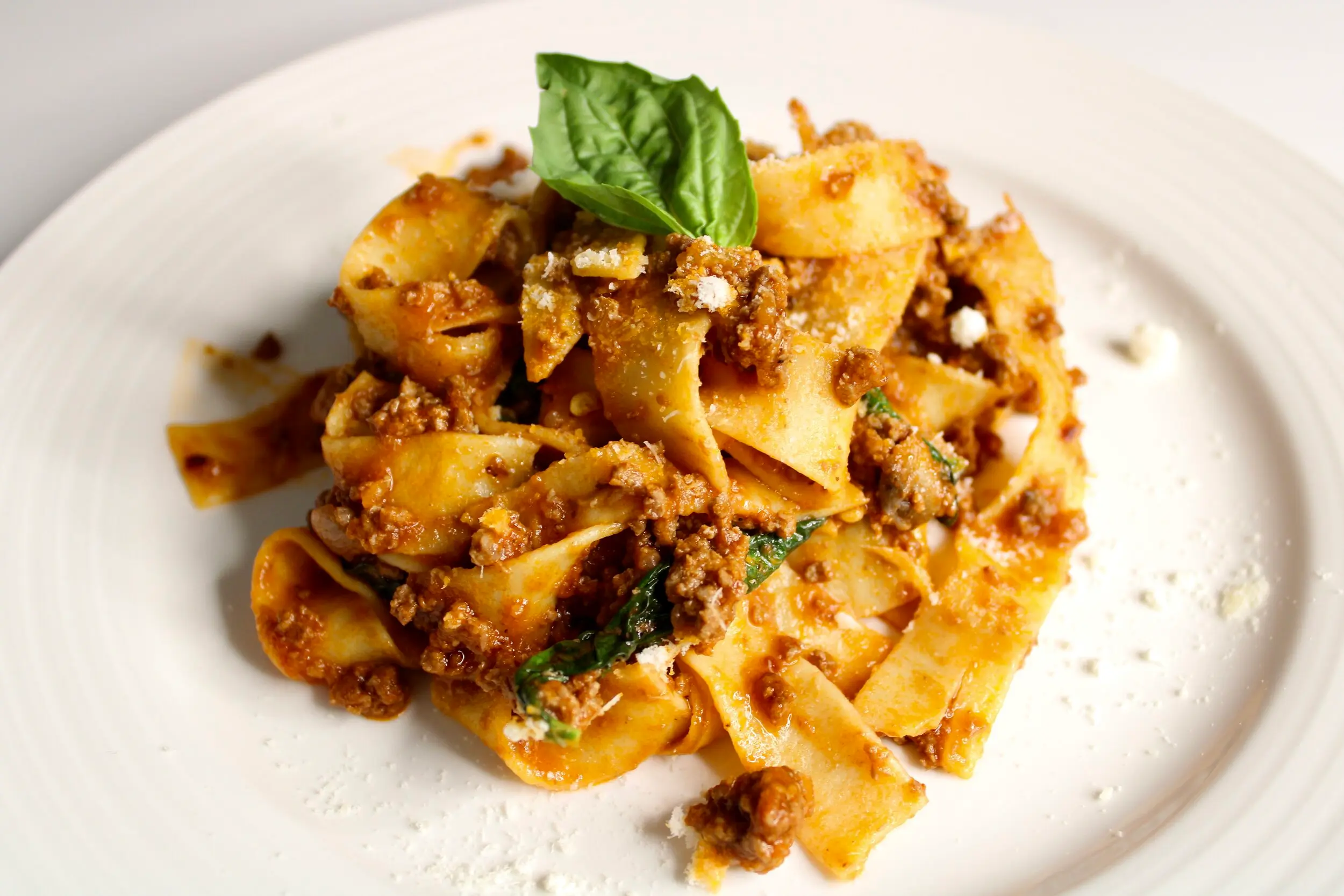 A cream coloured plate full of the infused pappardelle bolognese pasta with a basil leaf on top for garnish.