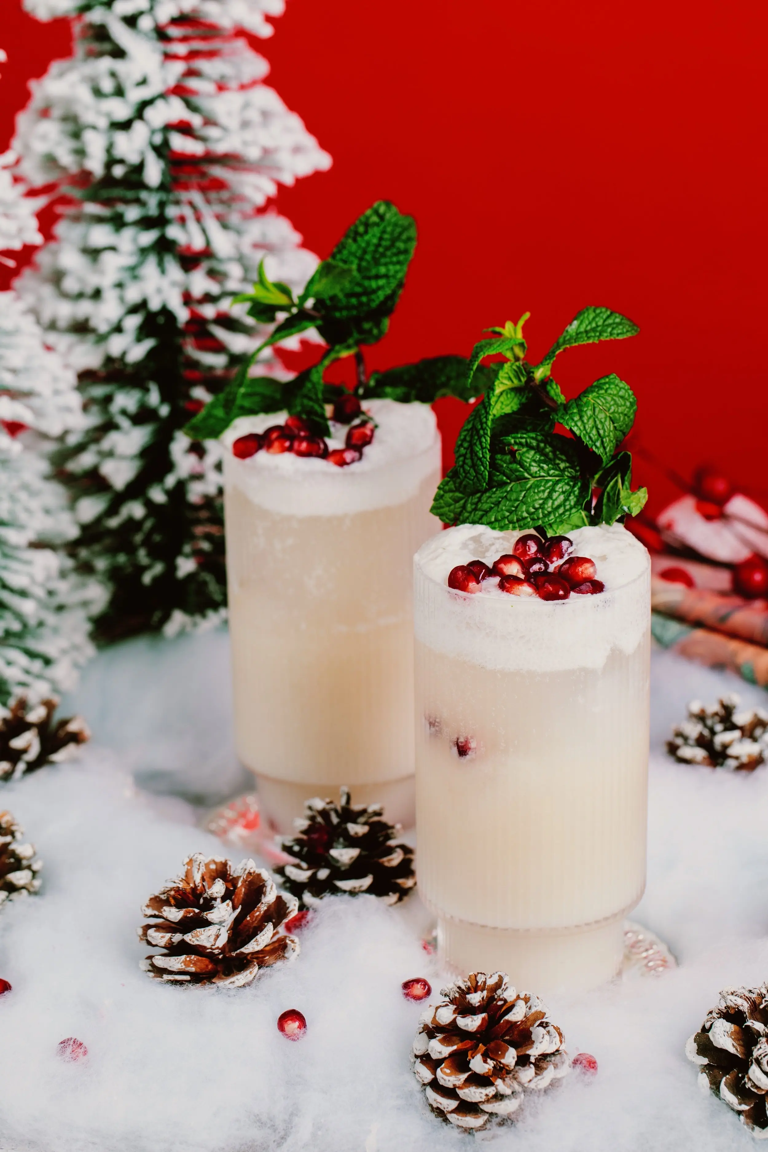 Two tall glasses full of a white/cear drink which is fizzy and overflowing at the top. It is garnished with pomegranate seeds and mint leaves and they sit on a bed of fluffy fake snow, surrounded by pinecones and Christmas trees.