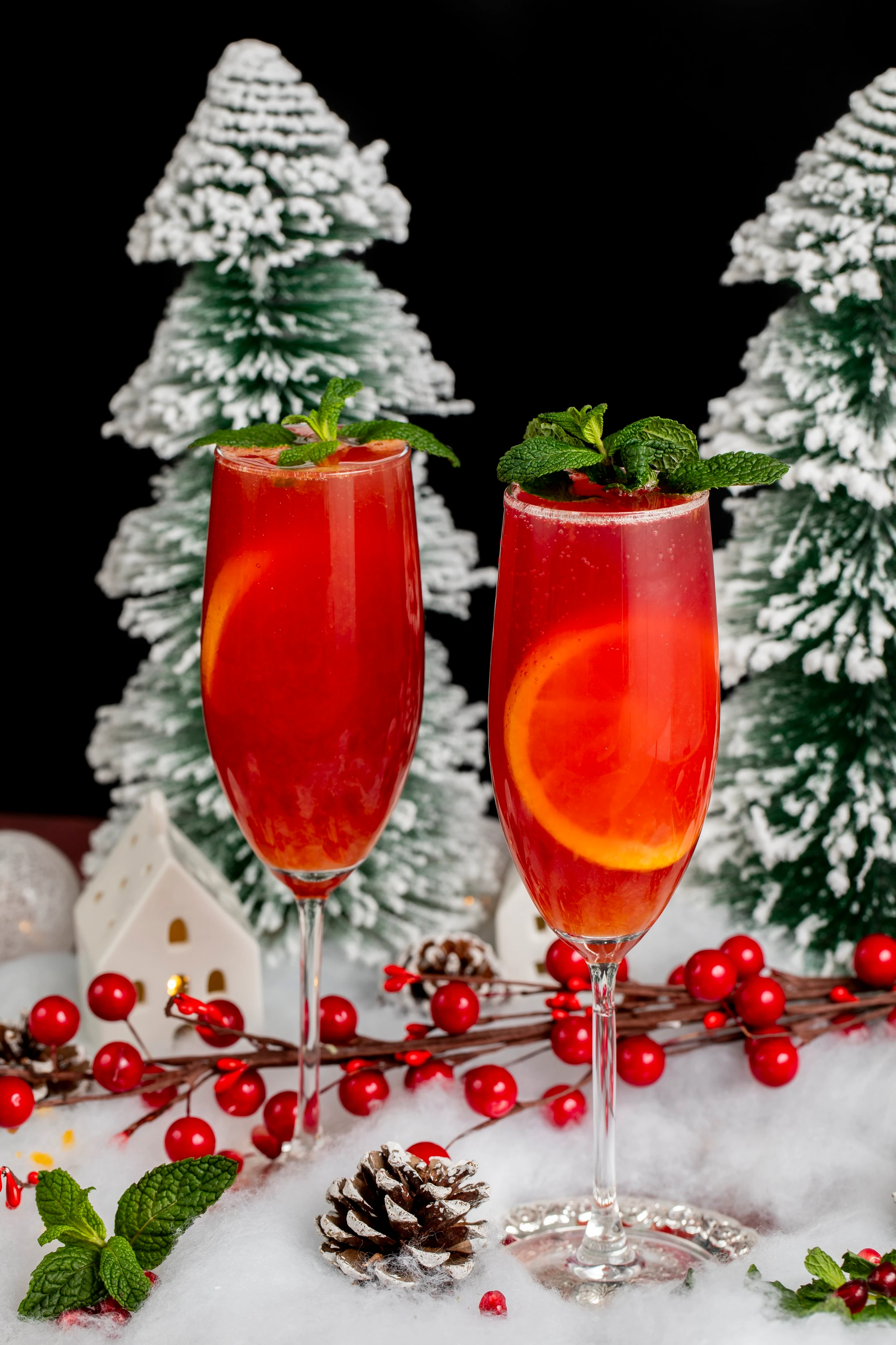 Two champagne flutes holding red cannabis infused mimosas garnished with mint leaves in front of a backdrop of frosted Christmas trees and berry garlands.