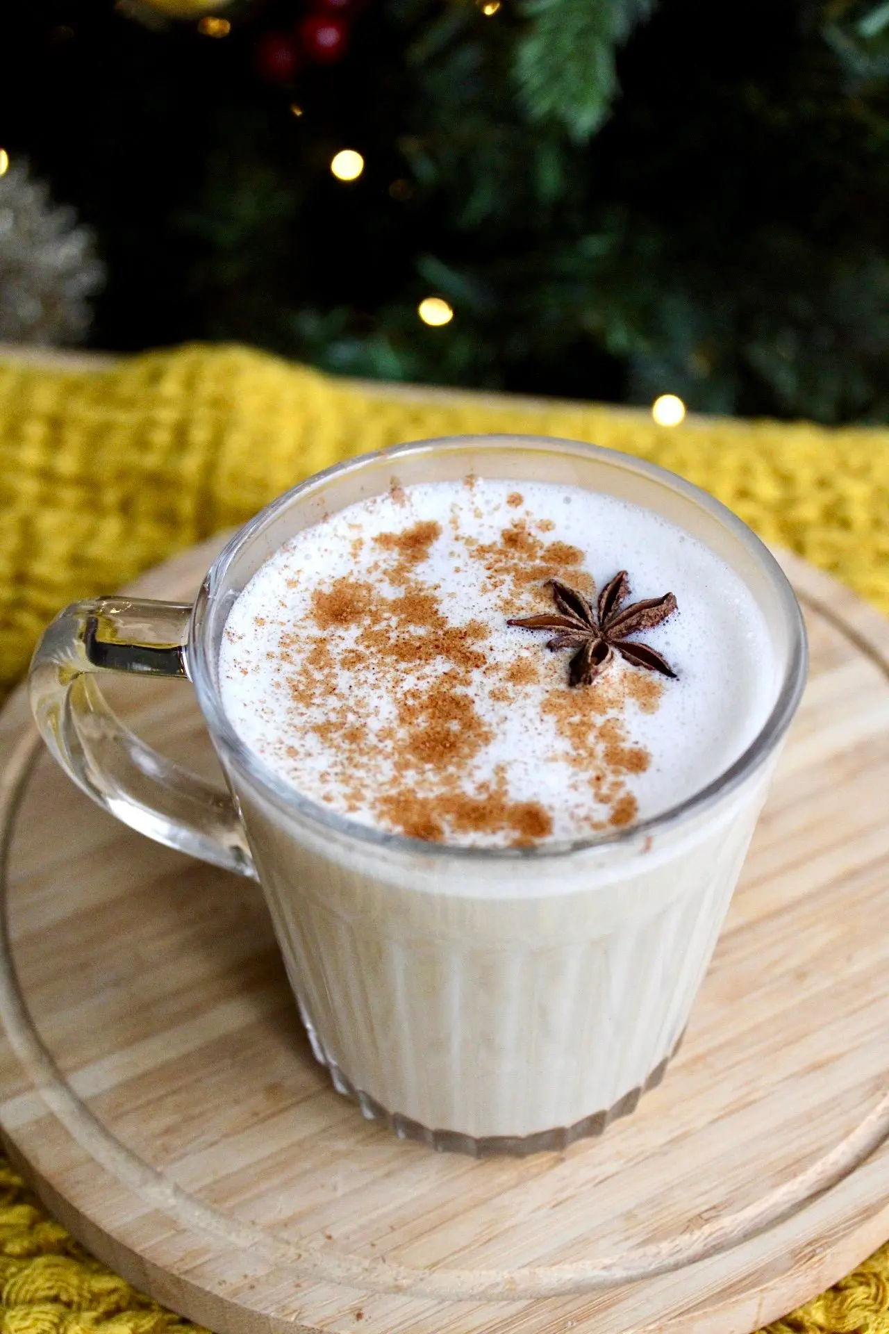 a clear glass mug filled to the talk with cream coloured egg nog. The background features a Christmas tree and a yellow blanket.