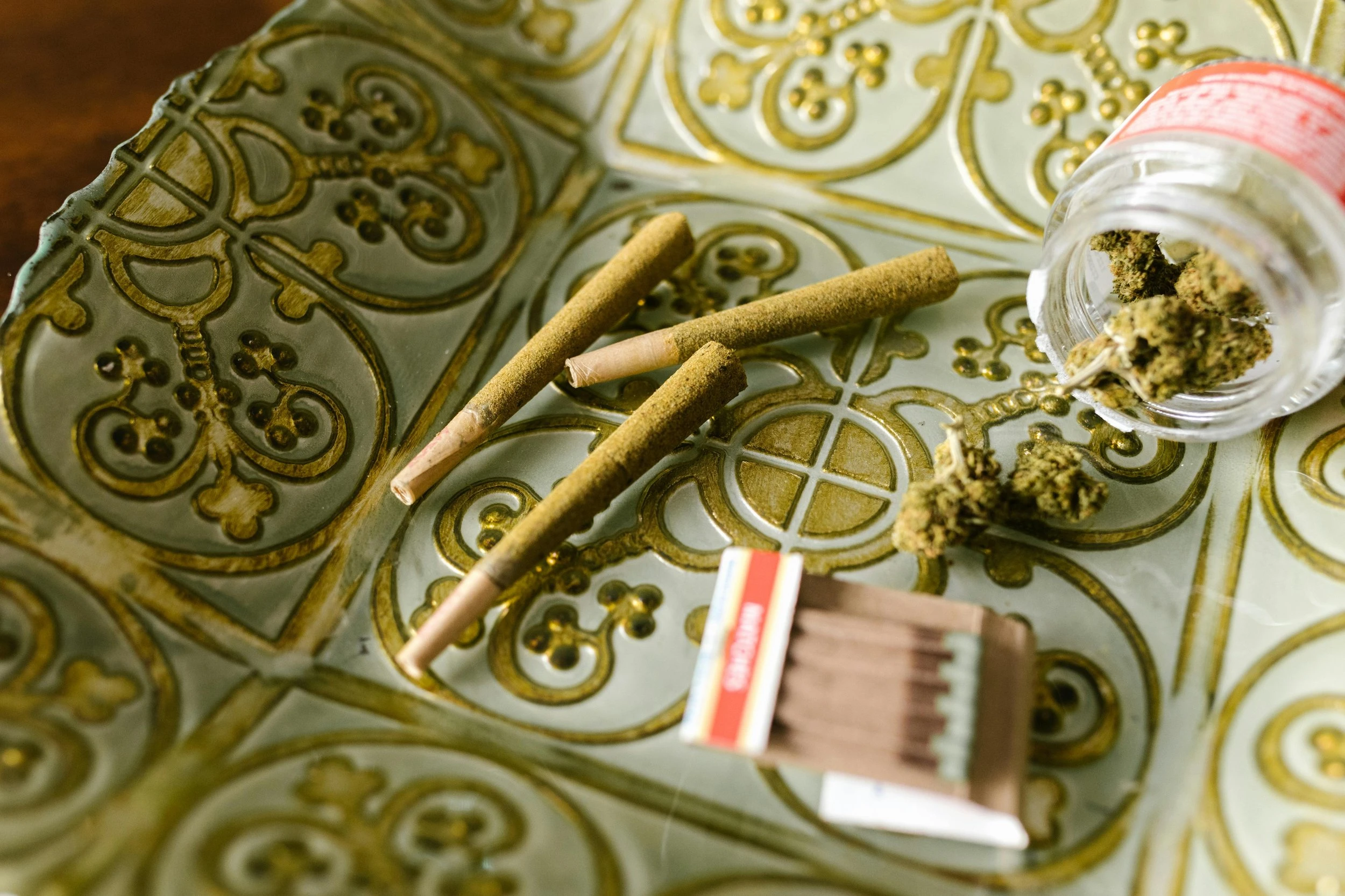 Three cannabis joints dipped in kief rest on an ornate green plate next to a set of matches and a jar of open, slightly spilled cannabis.