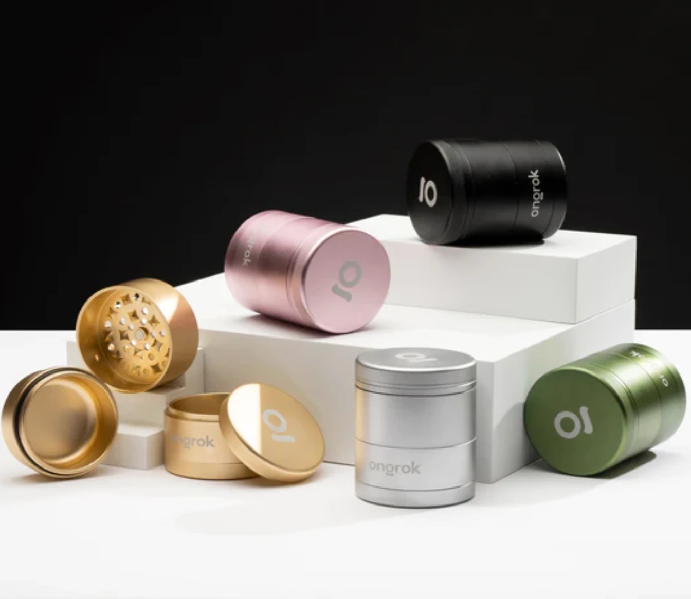 A pink, green, black, gold and silver Ongrok cannabis grinder rest on white blocks partially unattached to showcase the different pieces.