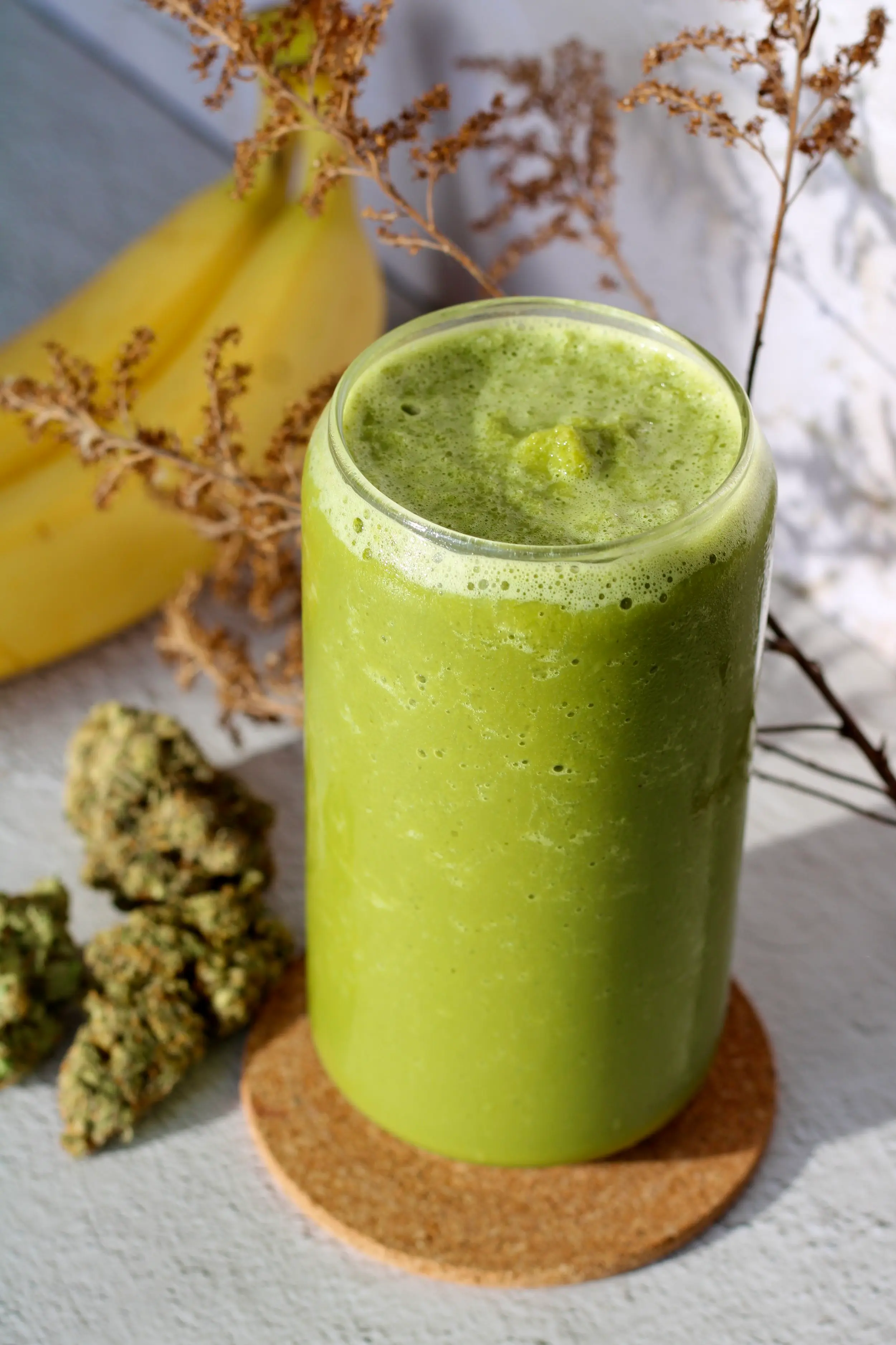 a glass full to the top of green matcha smoothie on a cork board coaster. Behind the glass is three different cannabis nuggets and two bananas.