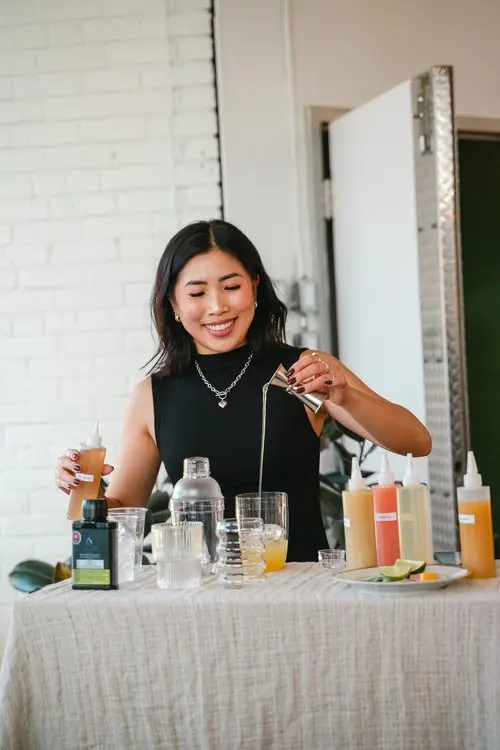 A photo of Anna The Cannabinista (a smiling woman with medium-length brown hair) pouring a liquid out of a jigger into a clear glass wth ice cubes in it. She is standing at a table and is surrounded by bottles of various liquids in various colours as well as other bar tools like a shaker. 