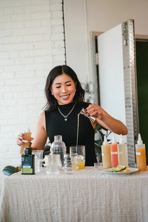 A photo of Anna The Cannabinista (a smiling woman with medium-length brown hair) pouring a liquid out of a jigger into a clear glass wth ice cubes in it. She is standing at a table and is surrounded by bottles of various liquids in various colours as well as other bar tools like a shaker. 