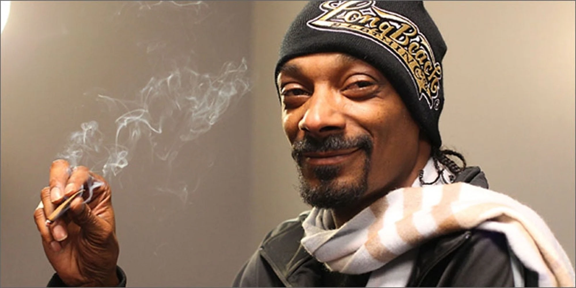 Snoop dog holding a lit cannabis joint and smiling at the camera with a beanie and scarf on. 