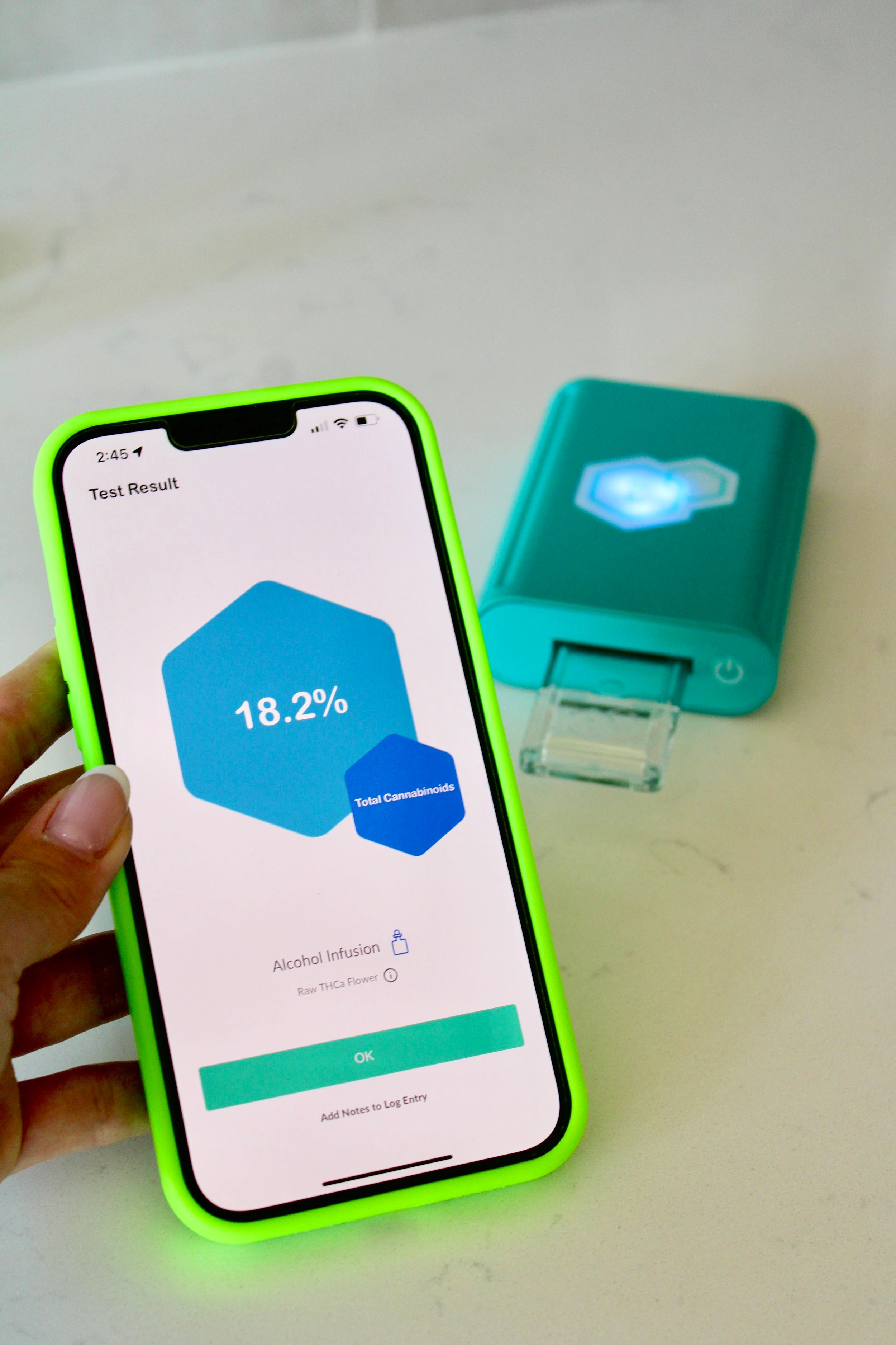 A blue t-check potency checker device rests on a table as an iPhone with a lime green phone case stands in front of it with a screen displaying the potency of a recently tested strain.