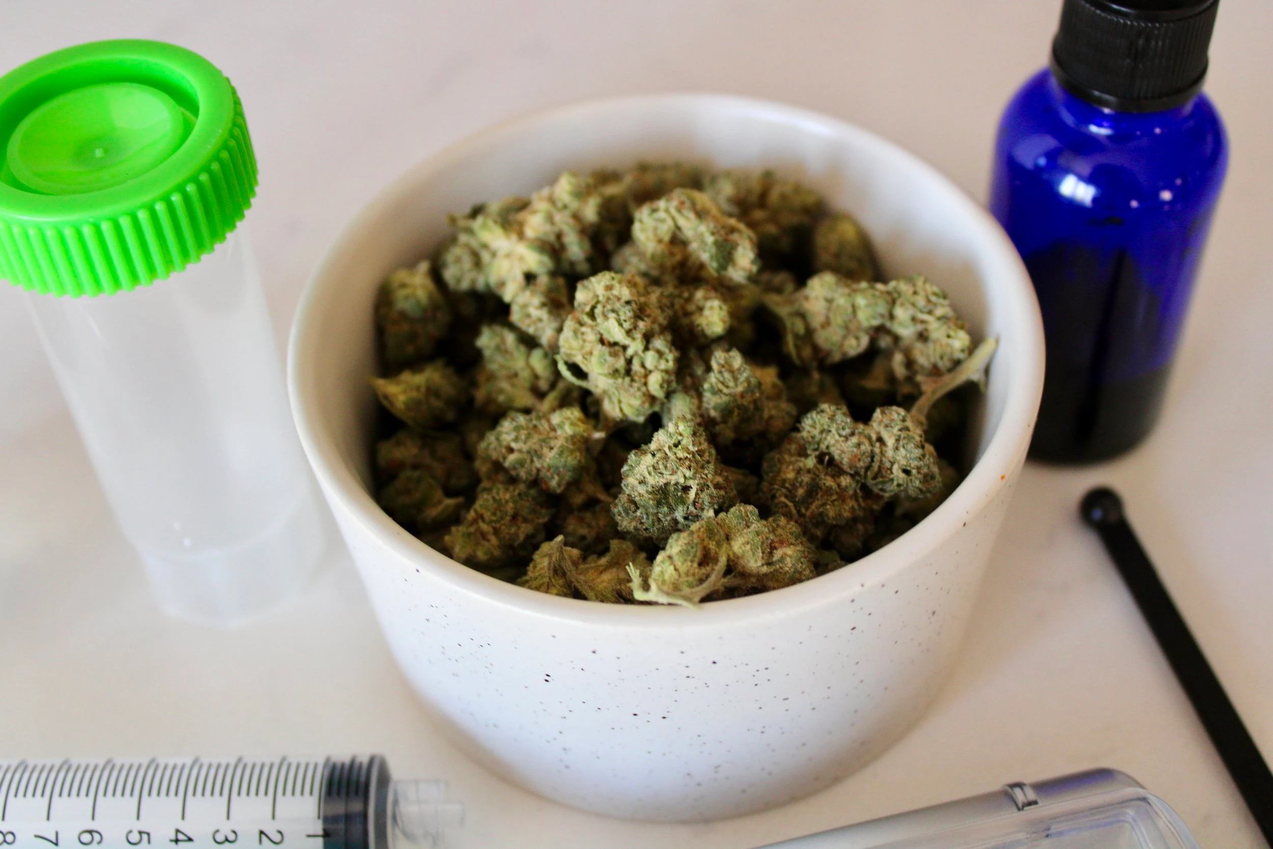 A large bowl of cannabis buds resting on a marble countertop next to an empty clear and green vial and a blue dropper bottle.