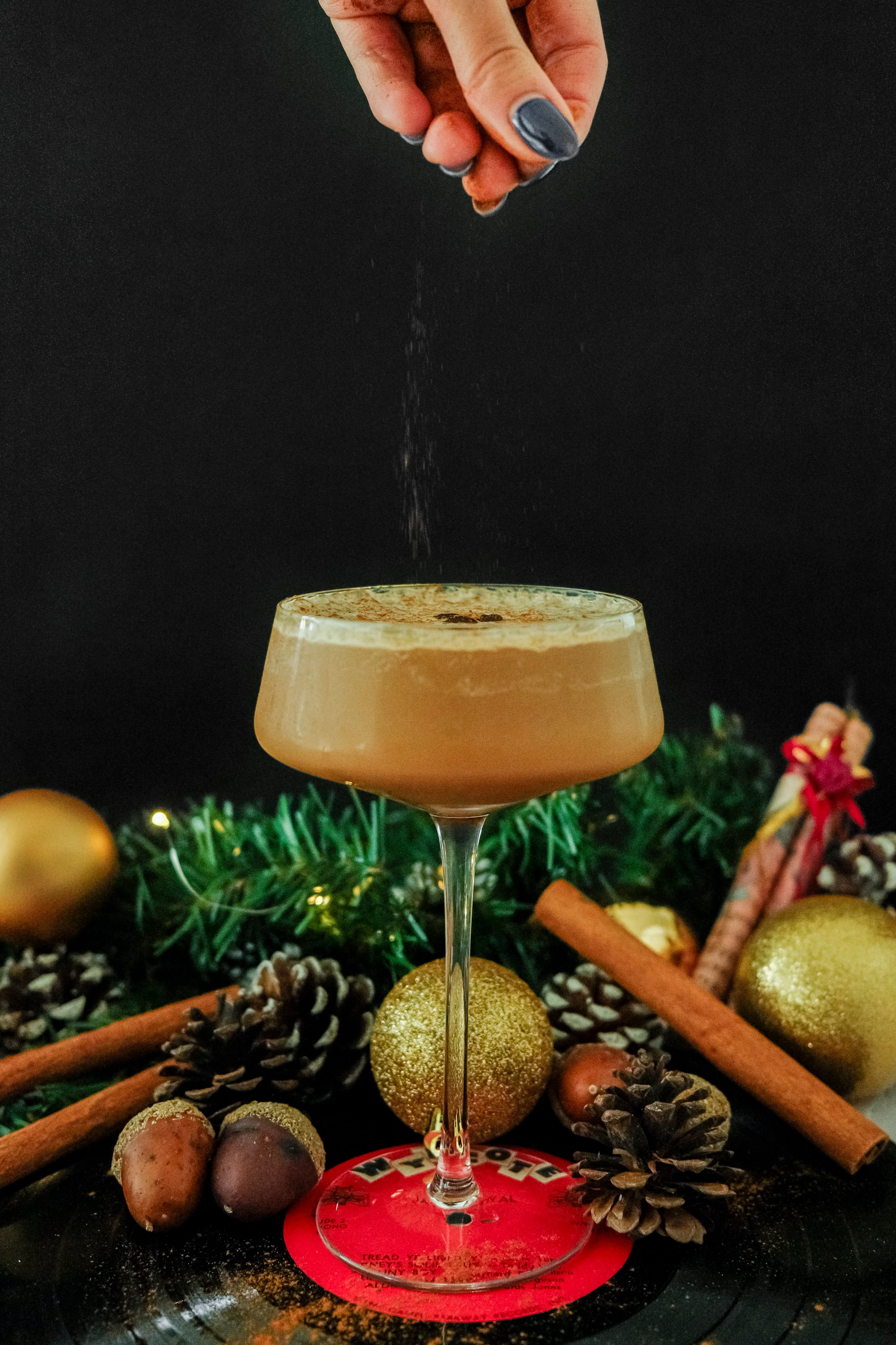 A long stemmed martini glass holding an espresso martini mocktail resting on a bright red coaster as a hand sprinkles cinnamon on the foamy top of the drink.