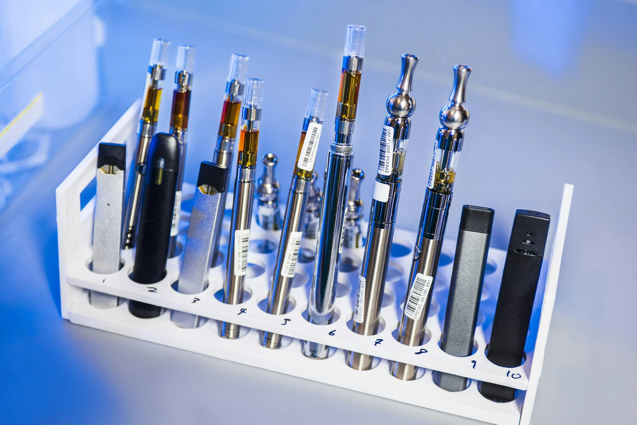 Many different kinds of vape pens stand in a white vape holder/display with a blue light shining on them in the background. 