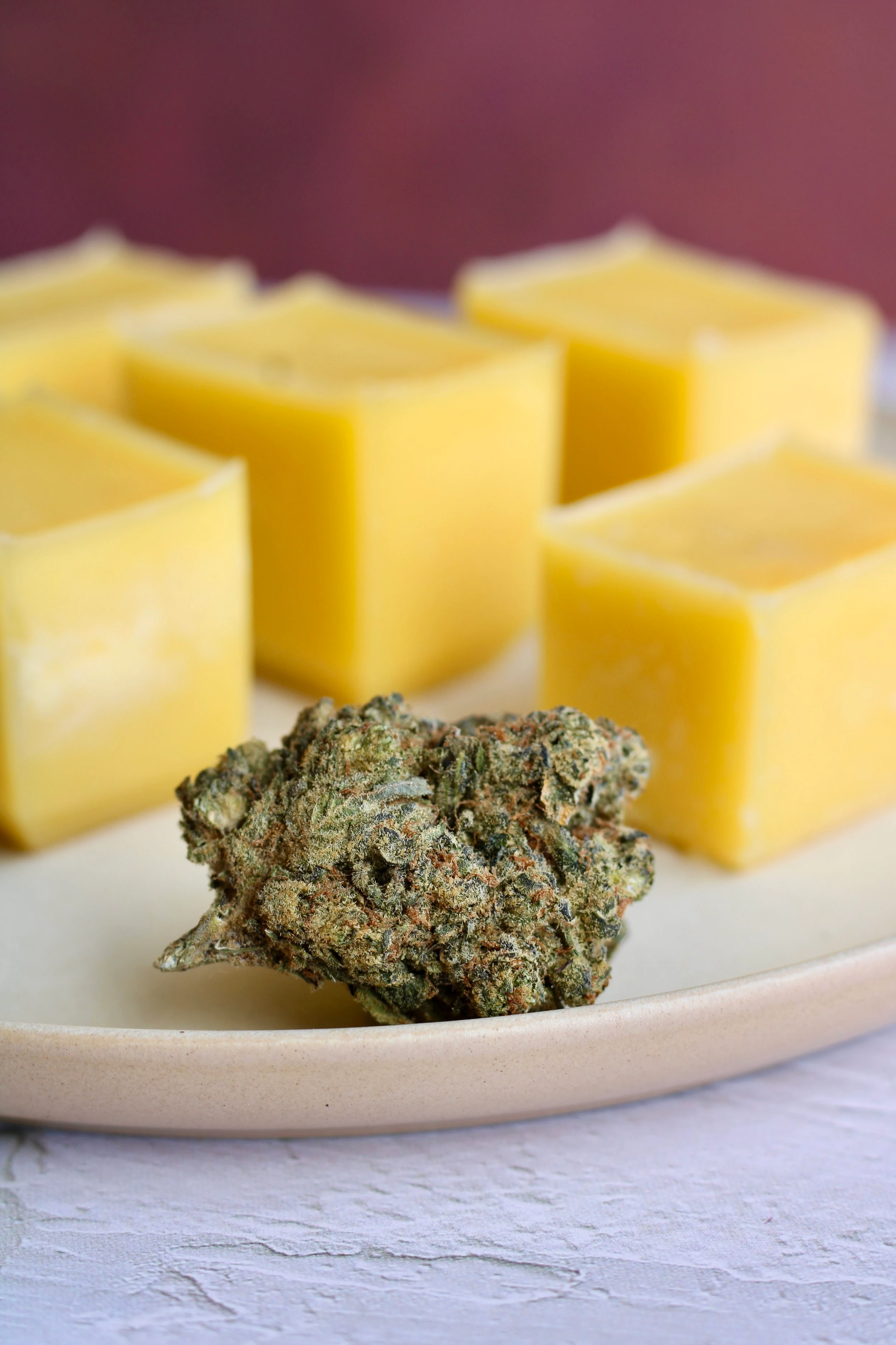 Five cubes of cannabis infused butter resting on a light beige plate, with a large cannabis nugget sitting in front of it.